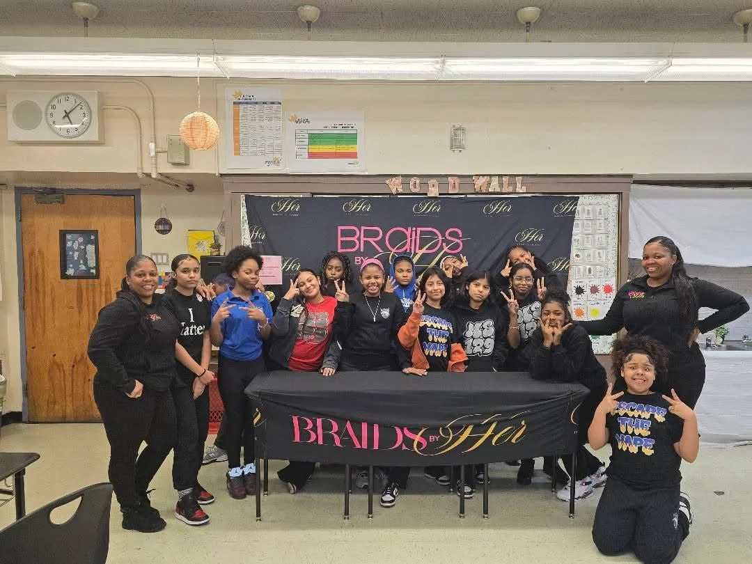 Group of young girls at a birthday party in a classroom or community room, standing behind a decorated table with a black tablecloth and banner reading 'Brides by Hia'. Some girls make peace signs and smiles, with a few posing playfully, and two wome