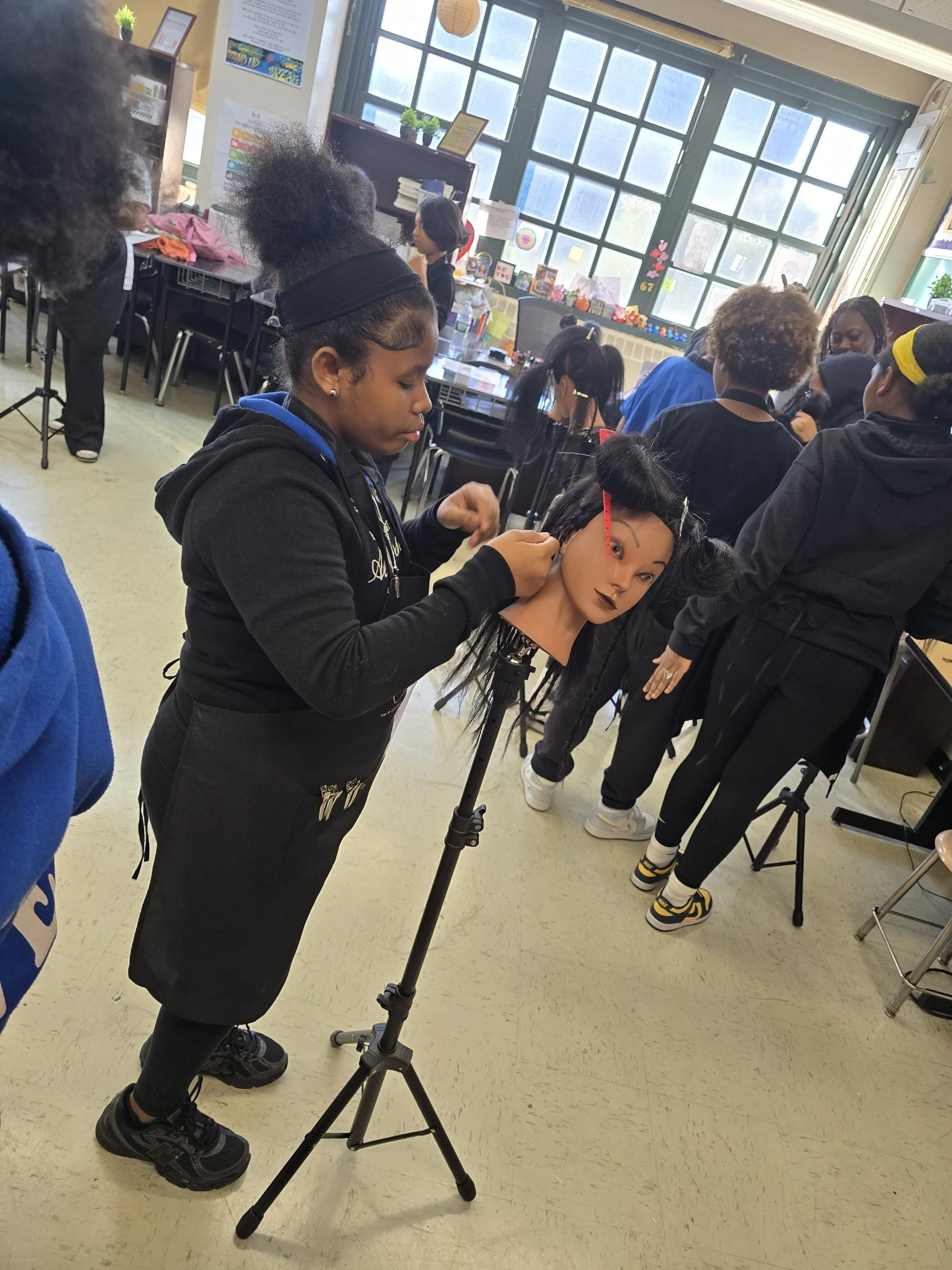 A young girl practicing hairstyling on a mannequin head in a classroom. Other students and teachers are present, some watching and others working. The classroom has large windows, desks, and various decorations.