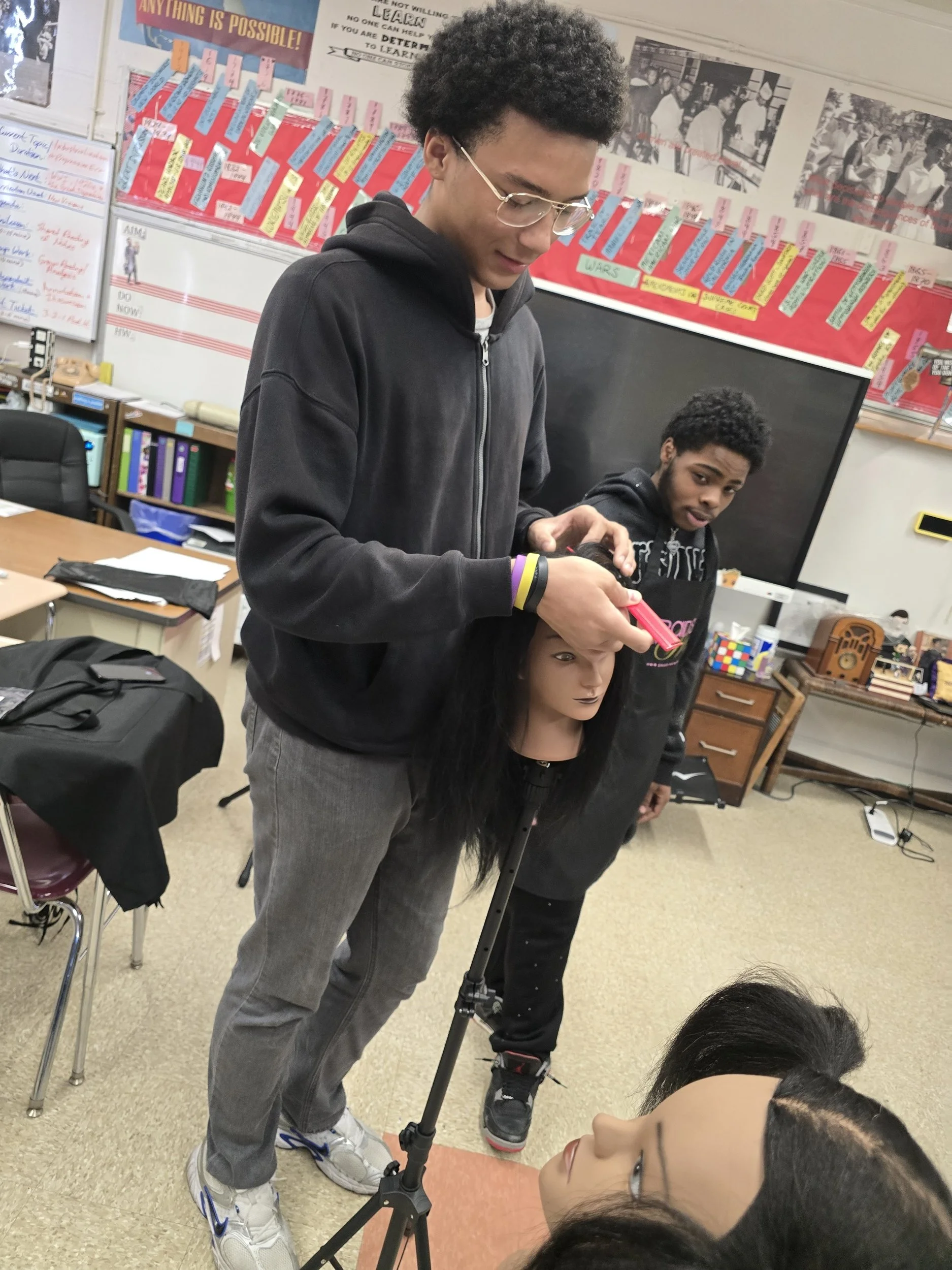 A classroom scene with a young man standing on a camera tripod, pointing it towards a girl who is lying on the floor. Another student is standing behind him, and there are desks, chairs, and classroom posters in the background.