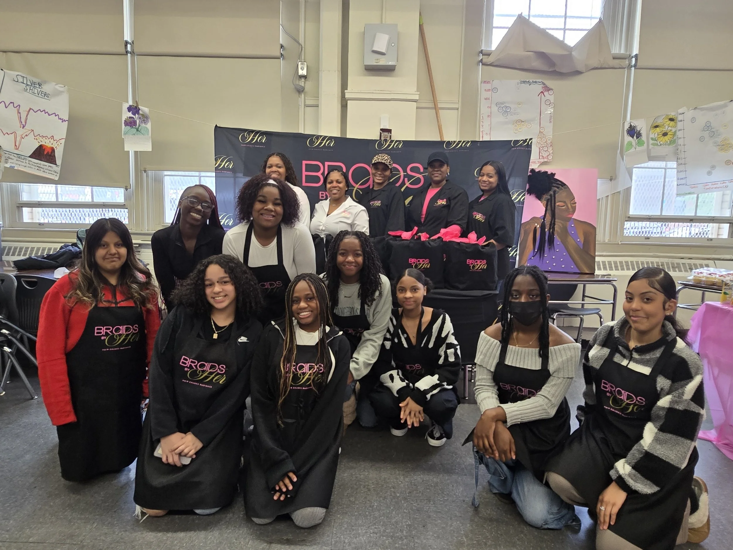 Group of women and girls posing indoors at an event, some wearing black aprons with pink text 'Bridal So.' There are partially visible posters on the walls, including drawings of volcanoes and flowers, and a large black banner with pink and white text.