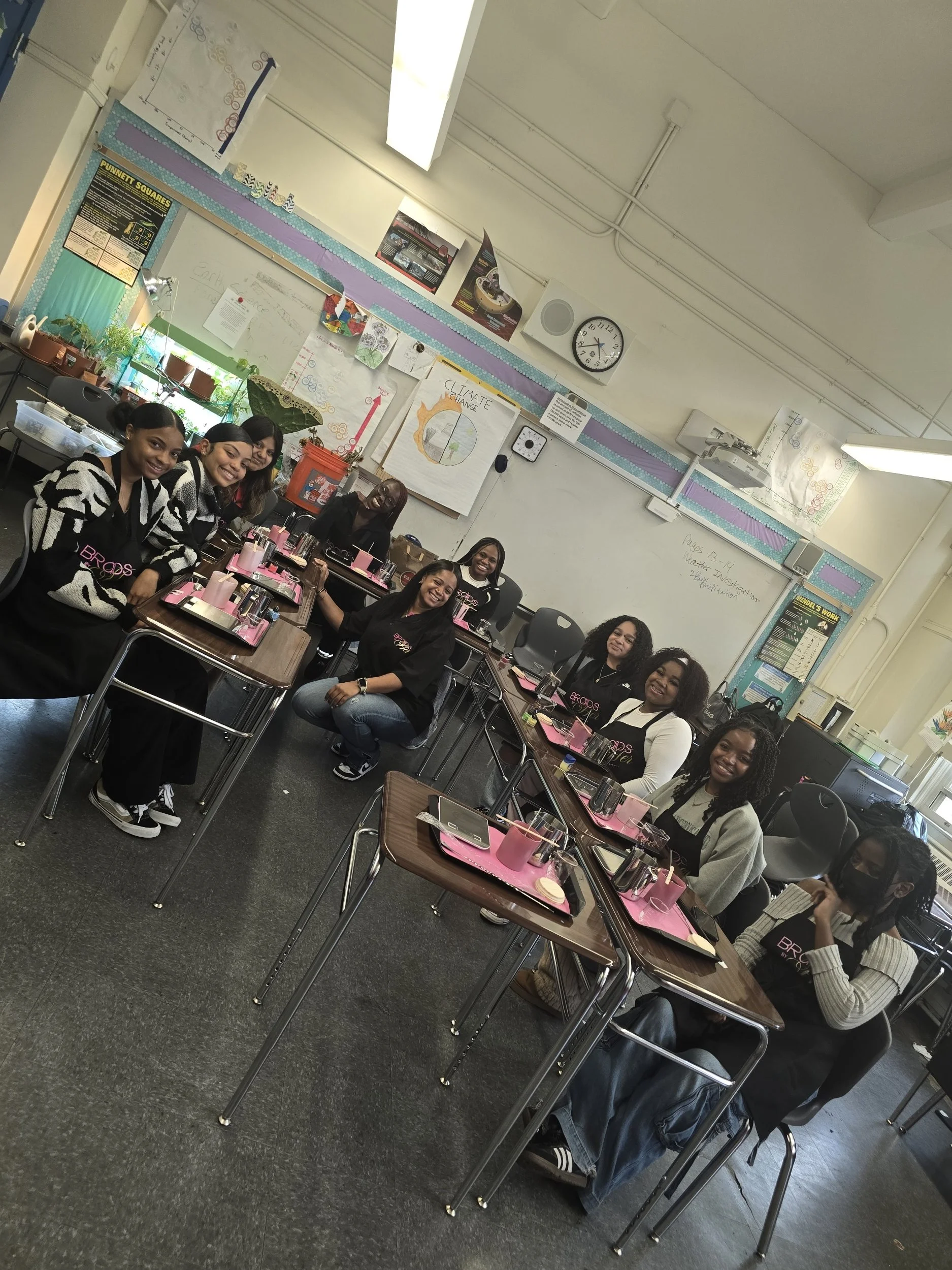 A group of women sitting at tables in a classroom or meeting room, smiling at the camera. The room has a whiteboard, clock, and various posters and charts on the walls. The tables are set with pink place mats and utensils, and there are potted plants