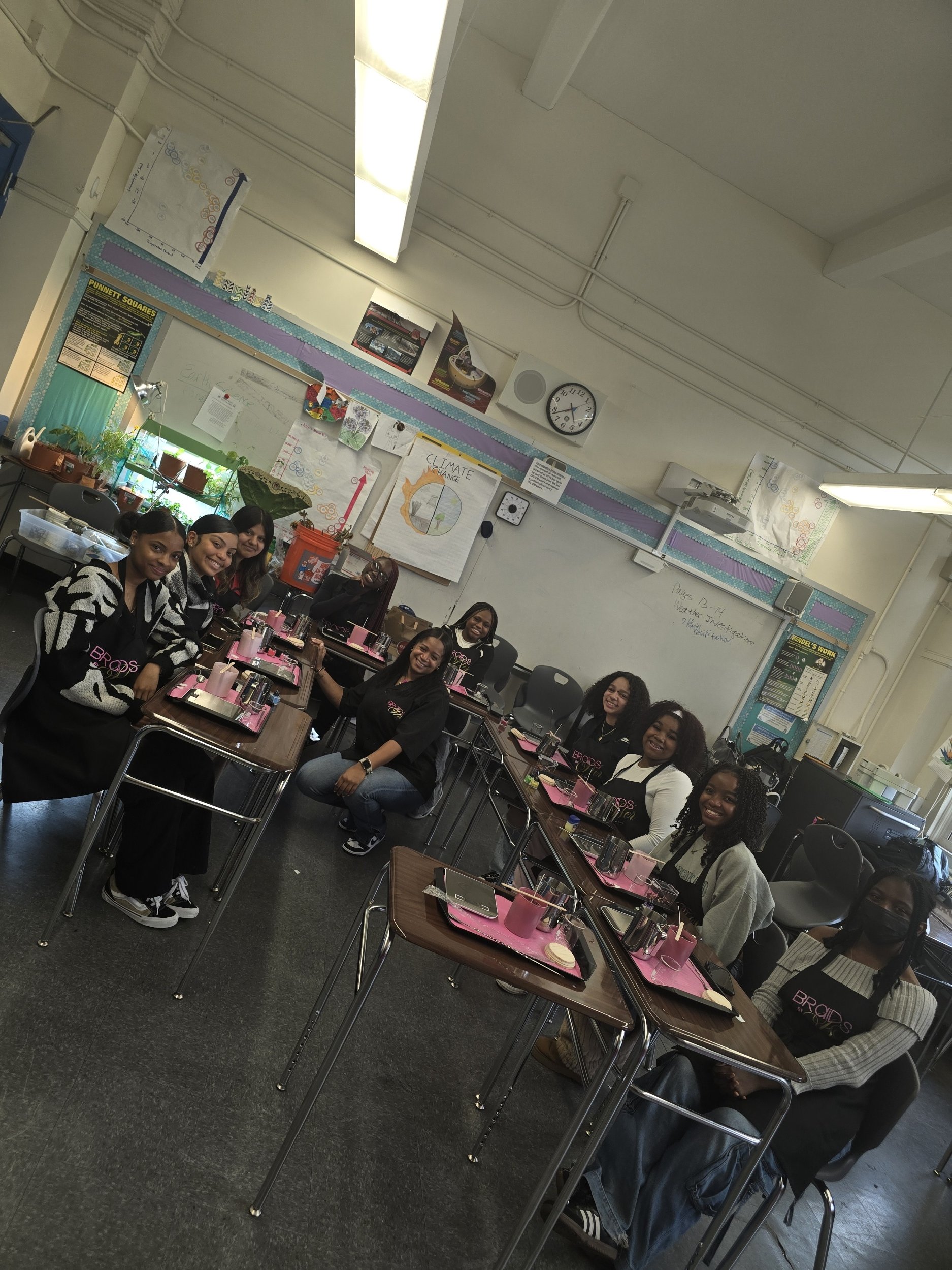 A group of smiling women sitting around tables in a classroom, prepared for a workshop or class, with plants and classroom decor in the background.