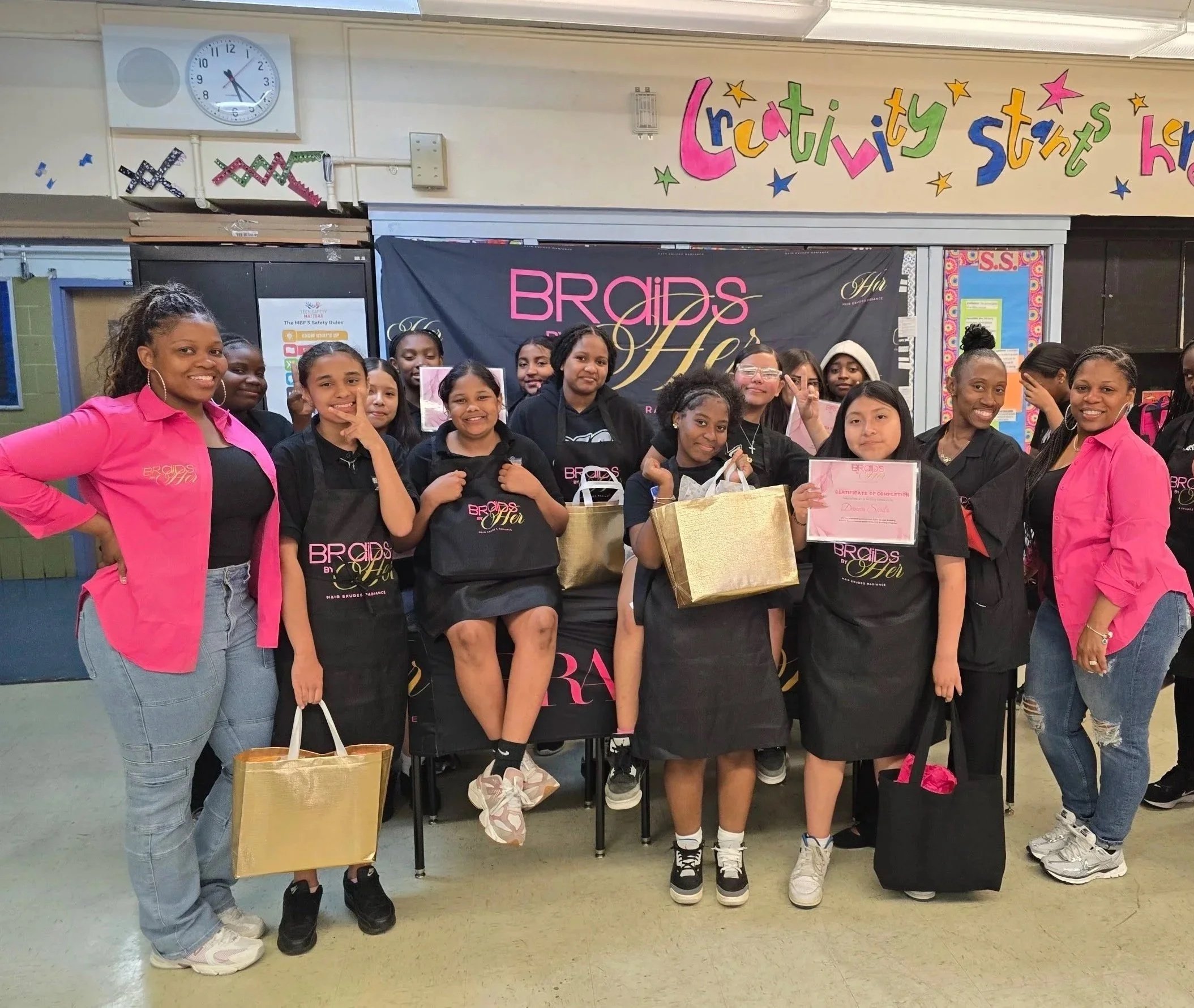Group of young girls and adult women posing at a group event in a school or community center, with colorful decorations and signs in the background, including a banner that says 'Braids & Her' and a sign with 'Creativity Starts Here'