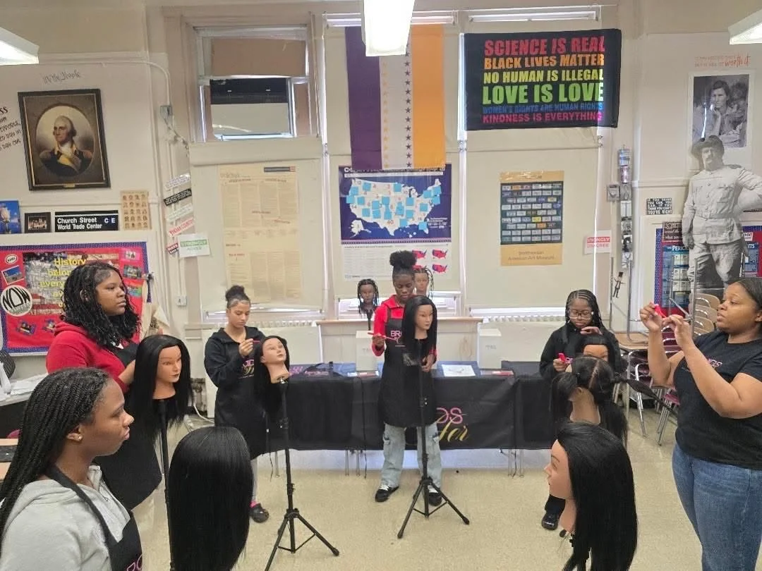 A group of young girls and women in a classroom, some mannequin heads and a woman taking a picture, with posters and a Liberty Bell picture on the wall.