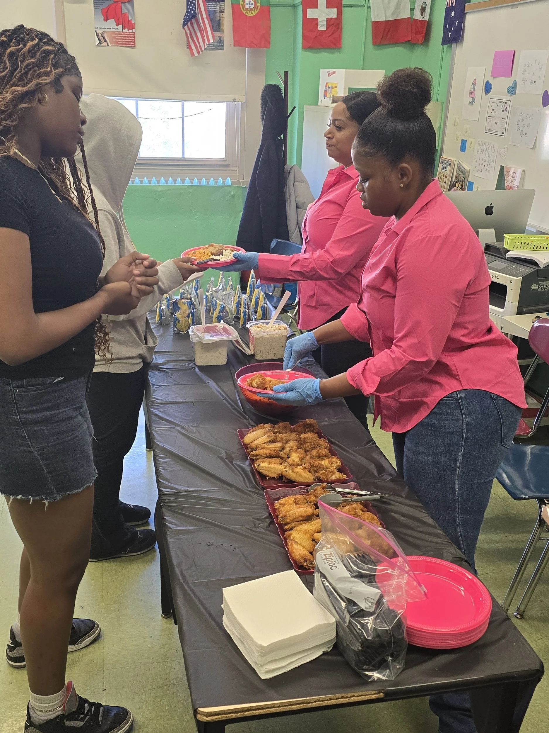 People serving and receiving food at a community event or gathering. Food items like fried chicken and other dishes are on the table, with some people wearing pink shirts helping serve. Flags and decorated walls are visible in the background.