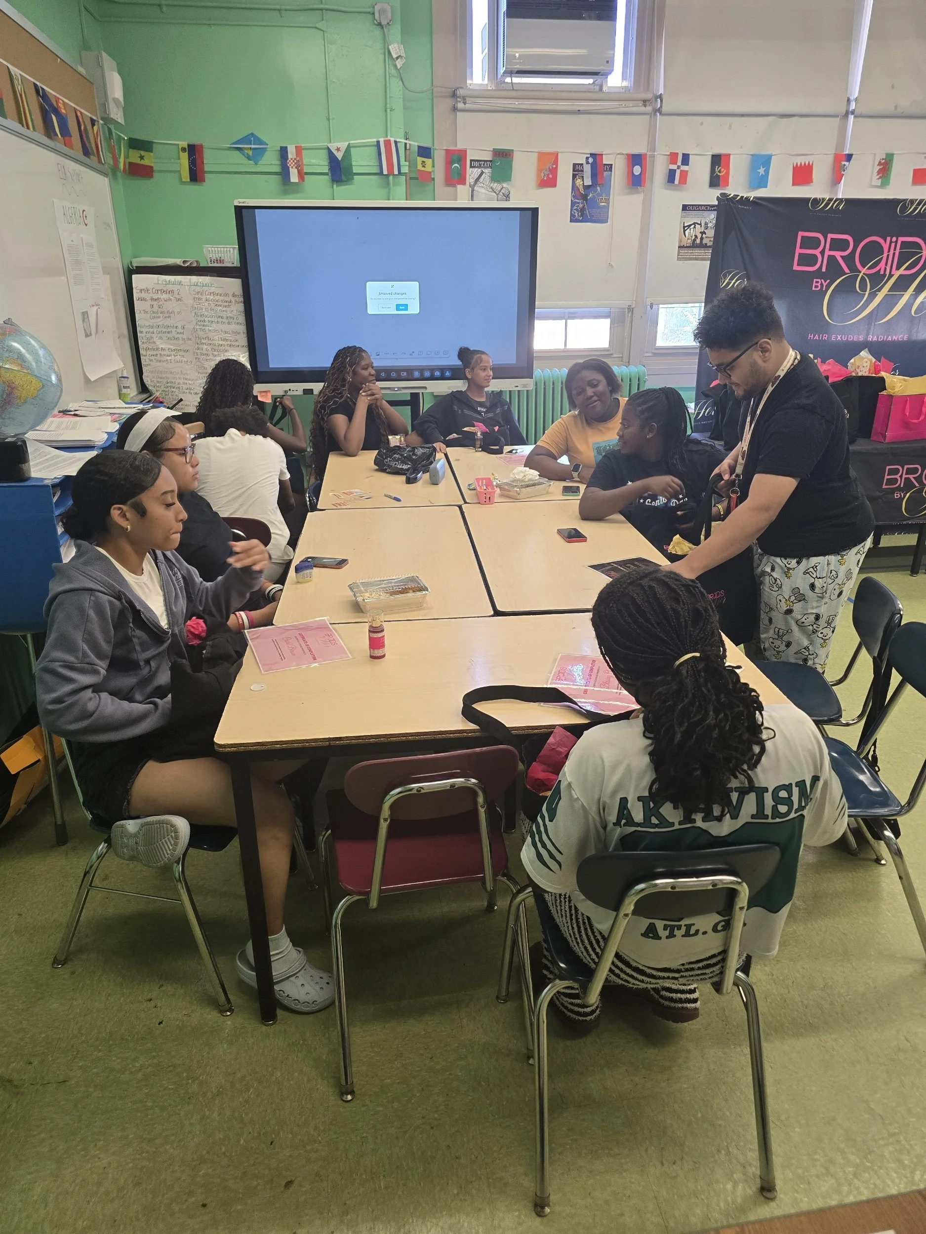 A classroom with students sitting at tables and a teacher standing. The room has a large screen, a world globe, and decorations including a string of international flags.