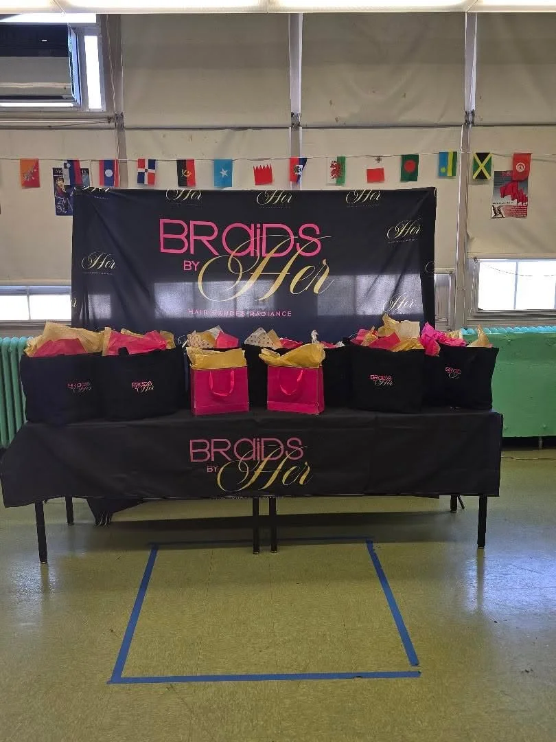 Table with gift bags and a banner for Braids by Ha, decorated with flags from various countries in the background, set up inside a room with a large window on the right.
