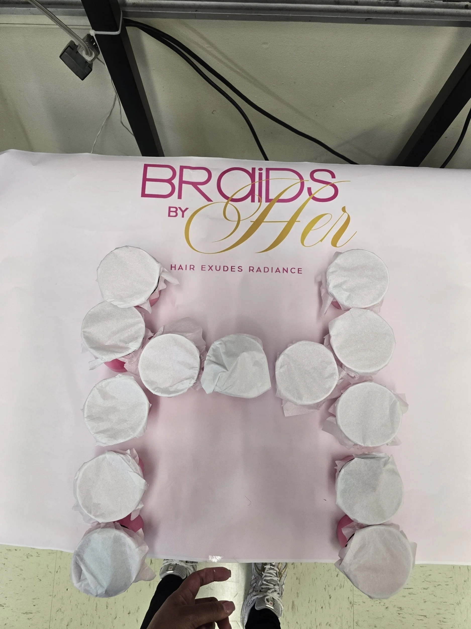 A person's hand holding a circular object wrapped in white tissue paper, placed on a pink box labeled 'Brads by Aeri' with the tagline 'Hair Exudes Radiance', surrounded by multiple white tissue paper-wrapped objects. The scene is in a room with a bl
