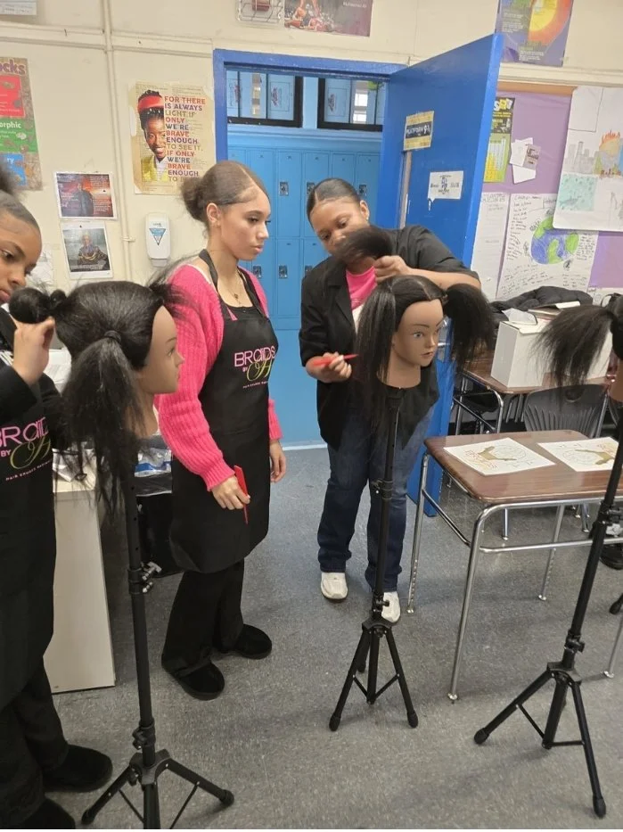 A group of students in a classroom practicing hairstyling on mannequin heads. Some students are holding tools like scissors and brushes, and one student is styling a mannequin's hair while others observe.