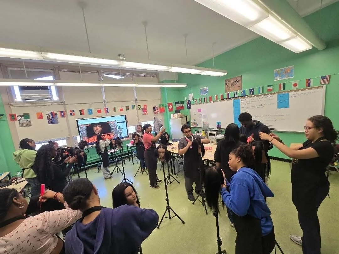 Students and teachers in a classroom preparing for a hair styling demonstration, with a large screen displaying a woman with curly hair.