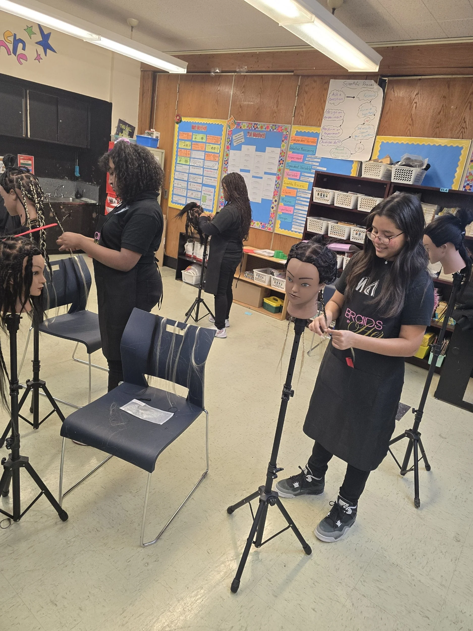 School classroom with students working on hair mannequin heads for hairstyling practice, with classroom posters and supplies visible in the background.
