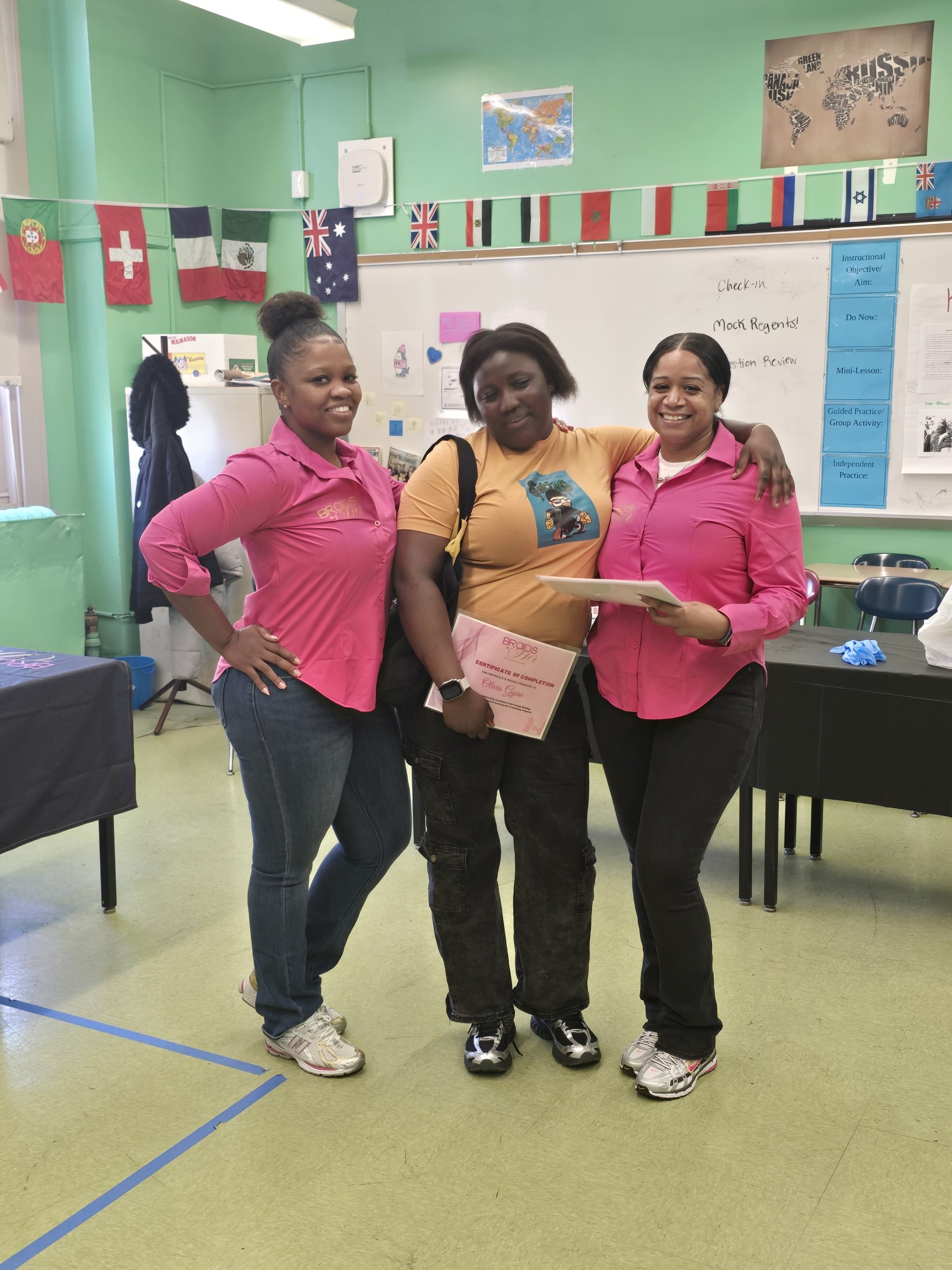 Three women standing together in a classroom, smiling and holding a certificate. Flags from various countries are hanging on a line and posters and a world map are on the walls behind them.