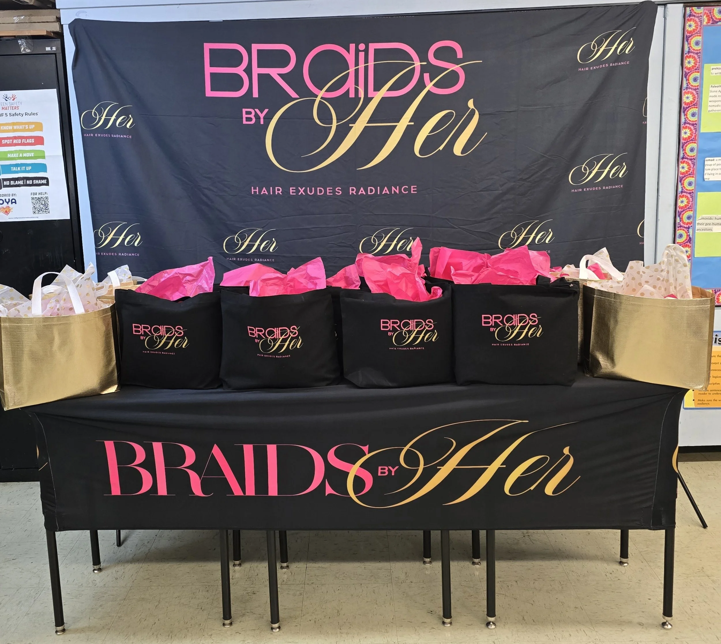A display table with black tablecloths featuring pink and gold lettering promoting a hair boutique named 'Braids by Her'. The background includes a large matching banner with the same branding, and various colorful gift bags with tissue paper are arr