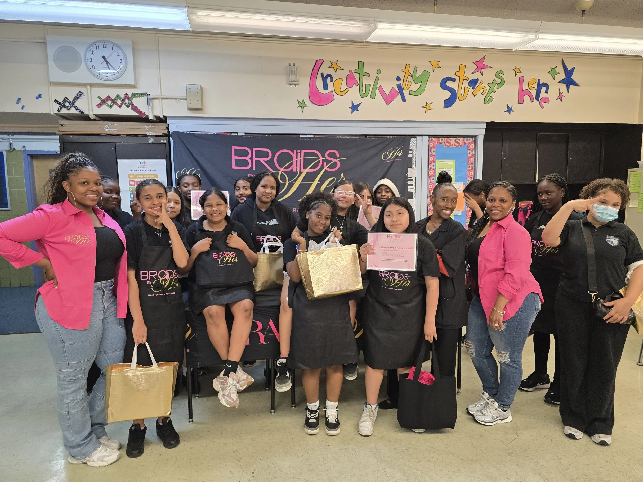 A group of girls and women smiling and posing for a photo in a classroom or school setting, with colorful signs and posters on the wall behind them and some holding gift bags and certificates.