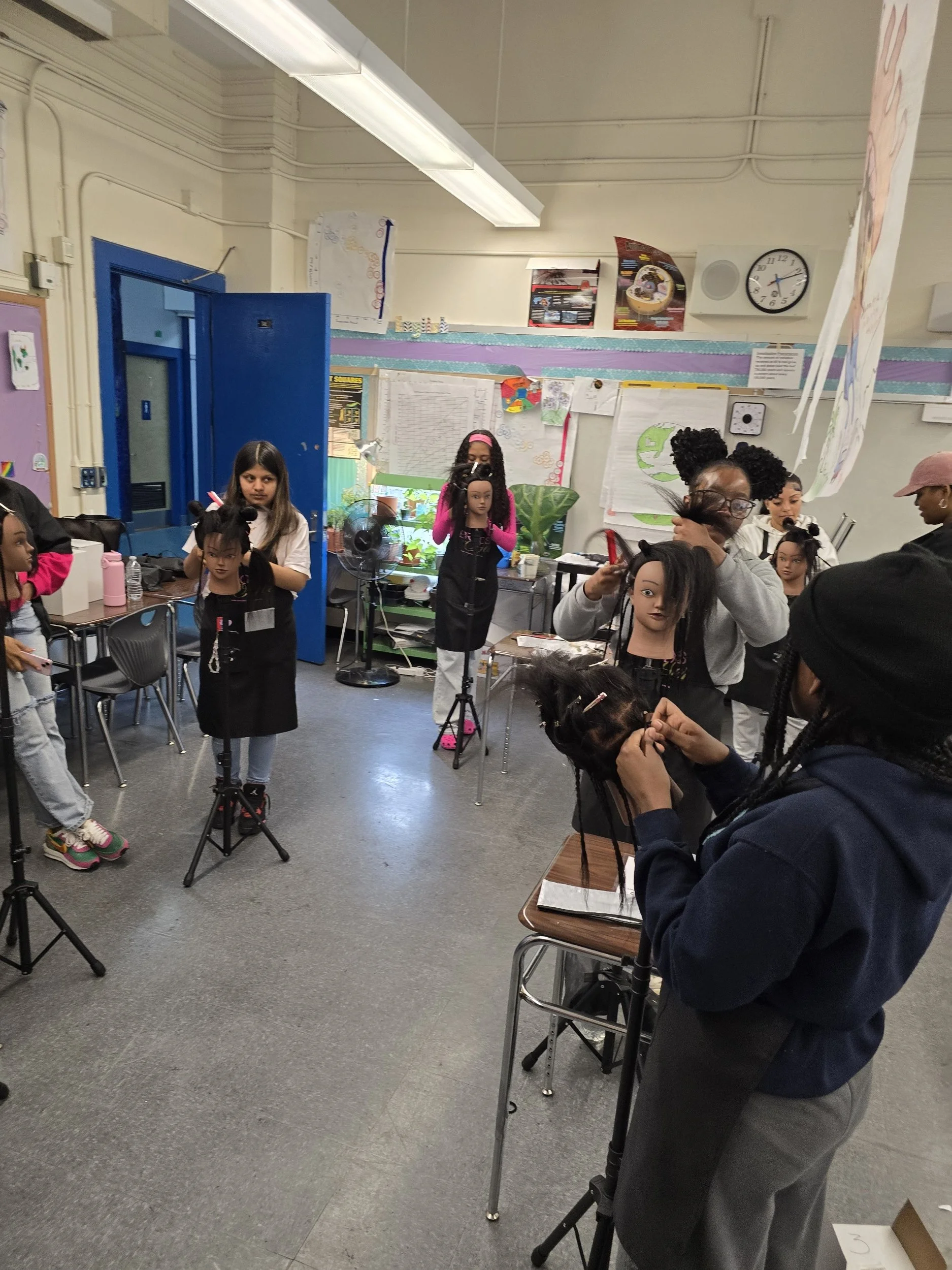 A classroom with people practicing hairstyling on mannequin heads, some standing and some sitting, with various posters, charts, and a clock on the walls.