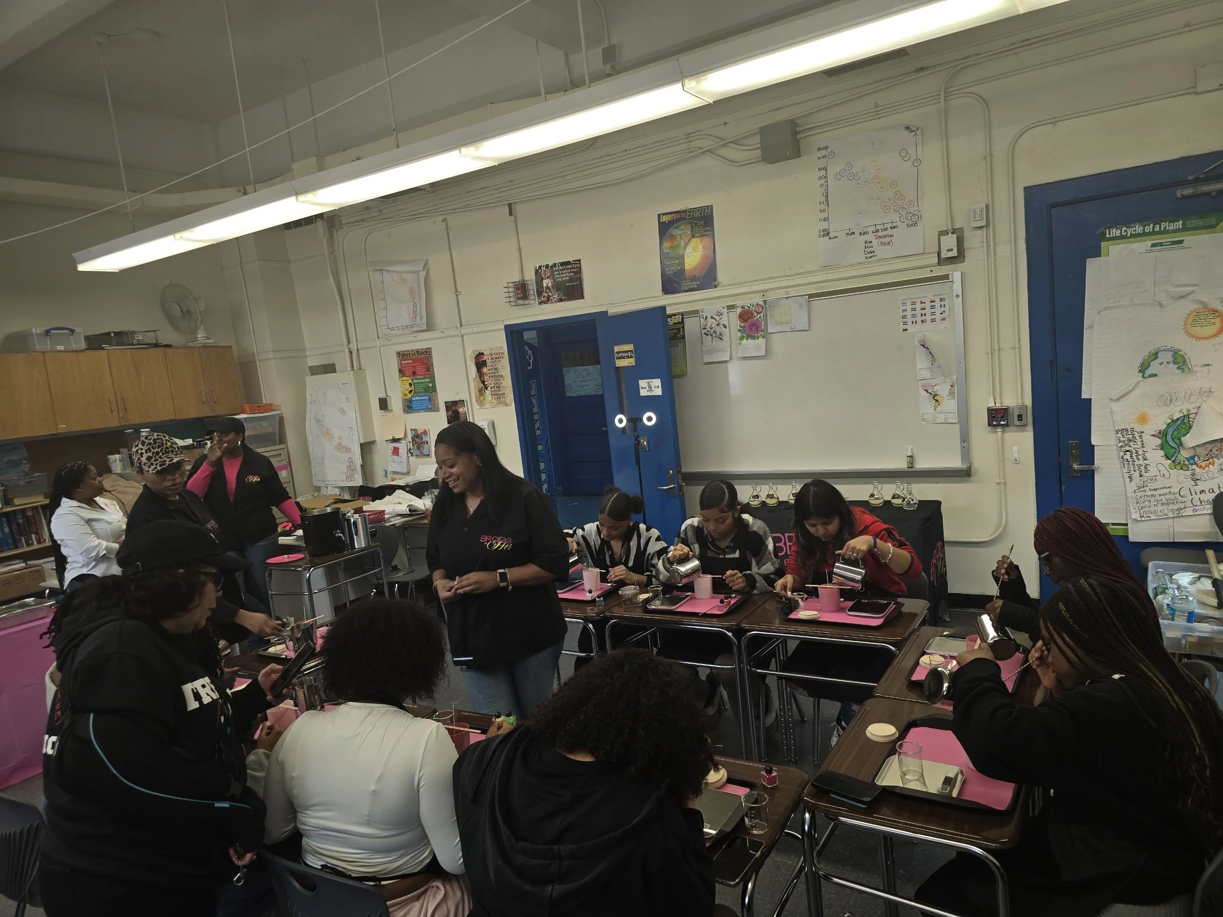 A classroom with several women sitting at tables, some working with small objects, and others standing and talking. The classroom has posters and charts on the walls, a blue door, and fluorescent lights overhead.