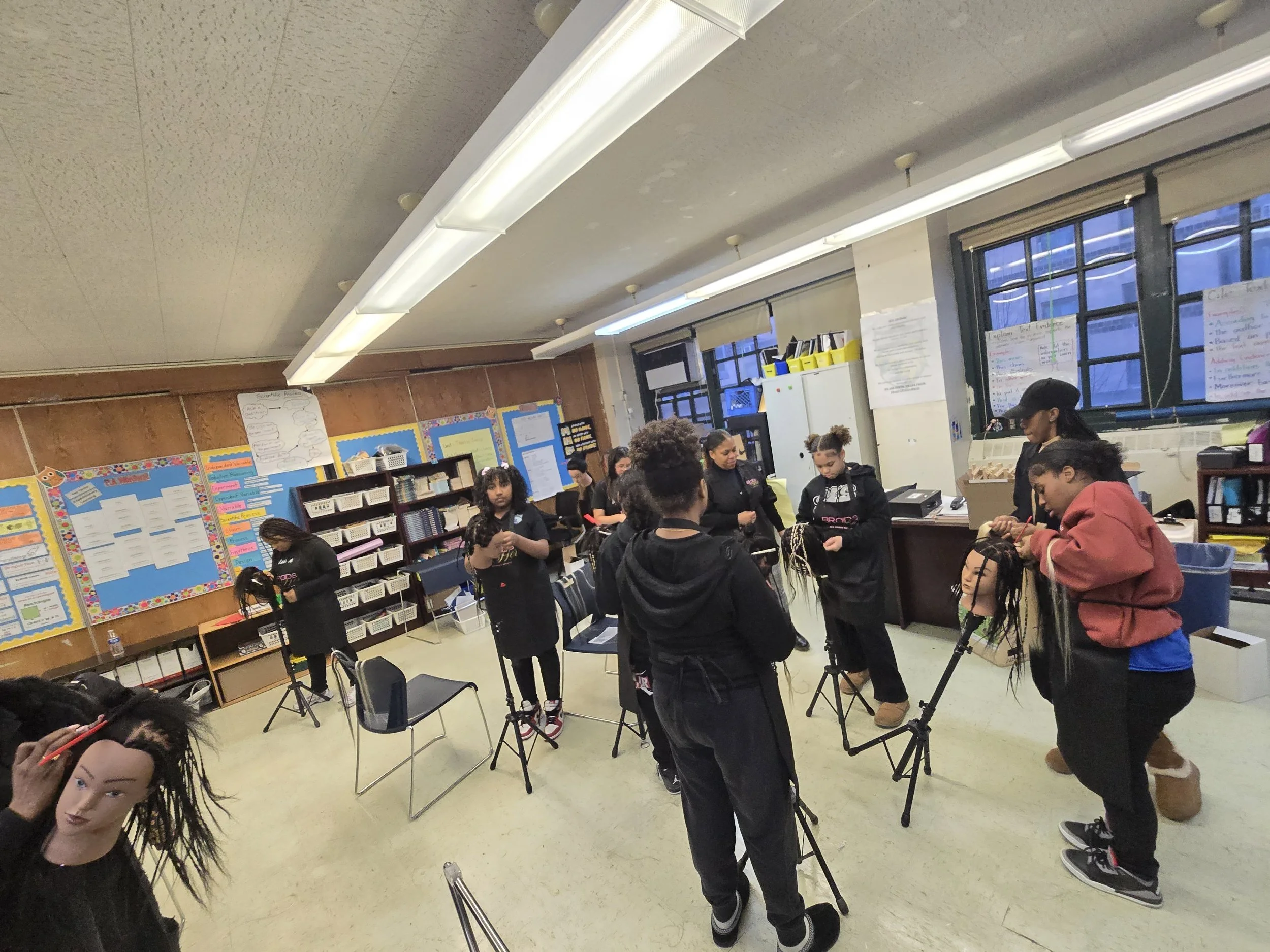 Classroom with students gathered around mannequin heads with braided hair, shelves with books and bins, colorful posters on bulletin boards, ceiling lights, and large windows.