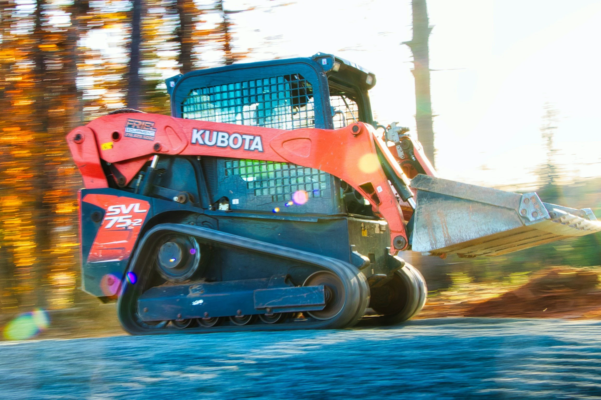 A red and black Kubota skid-steer loader moving through a wooded area with trees in the background, with sunlight creating a bright glow.