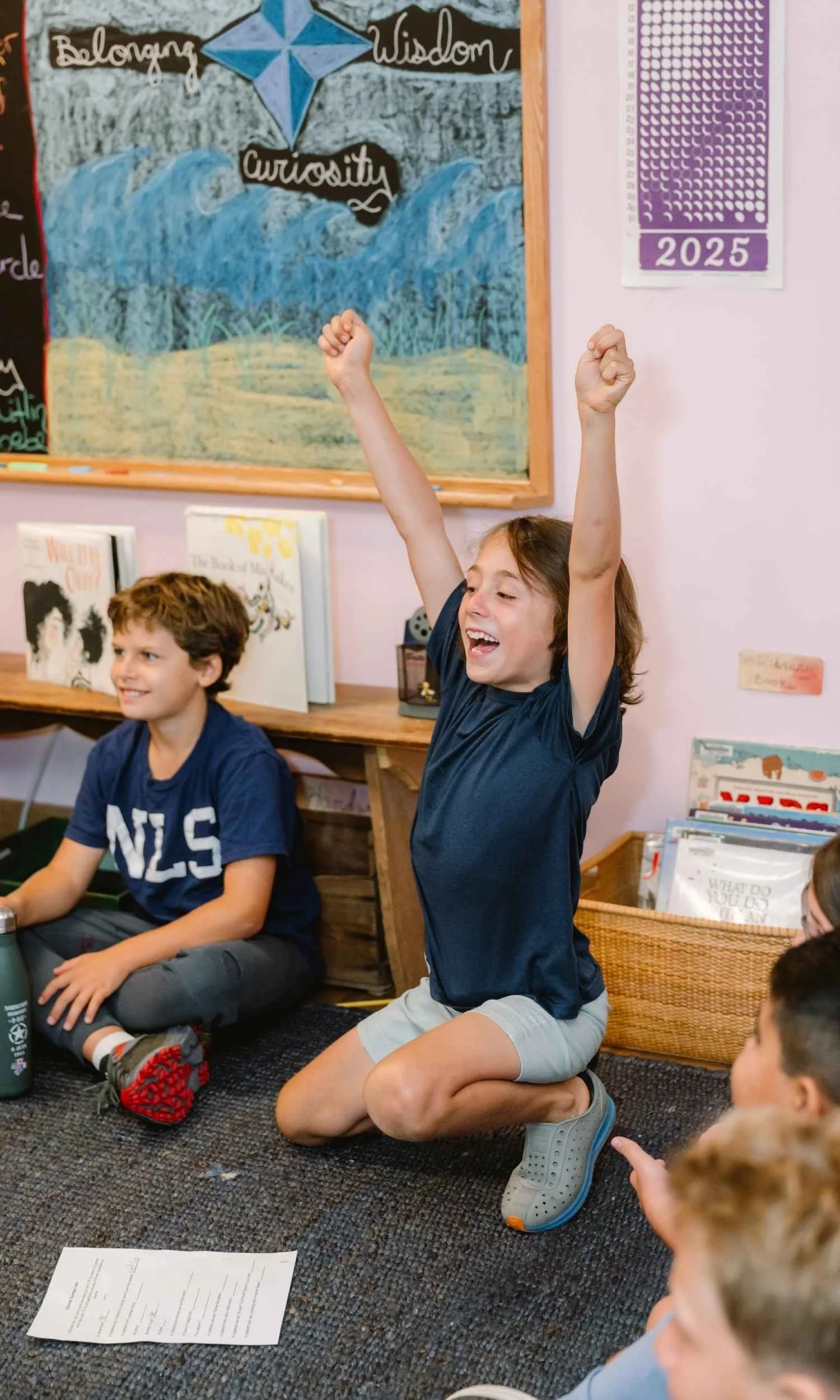 Two Nantucket Lighthouse School students sit together on a classroom floor, one raising his hands enthusiastically while the other smiles, in a welcoming learning space featuring a colorful chalkboard celebrating belonging, wisdom, and curiosity.