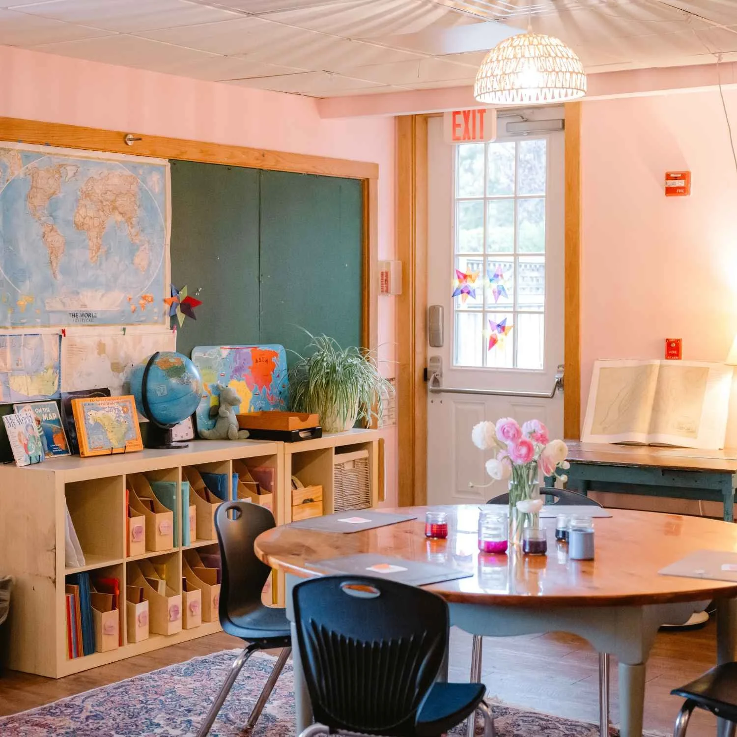 A bright Nantucket Lighthouse School classroom featuring wooden tables and chairs, educational posters, maps, books, and a globe, with natural sunlight filling the welcoming learning space.