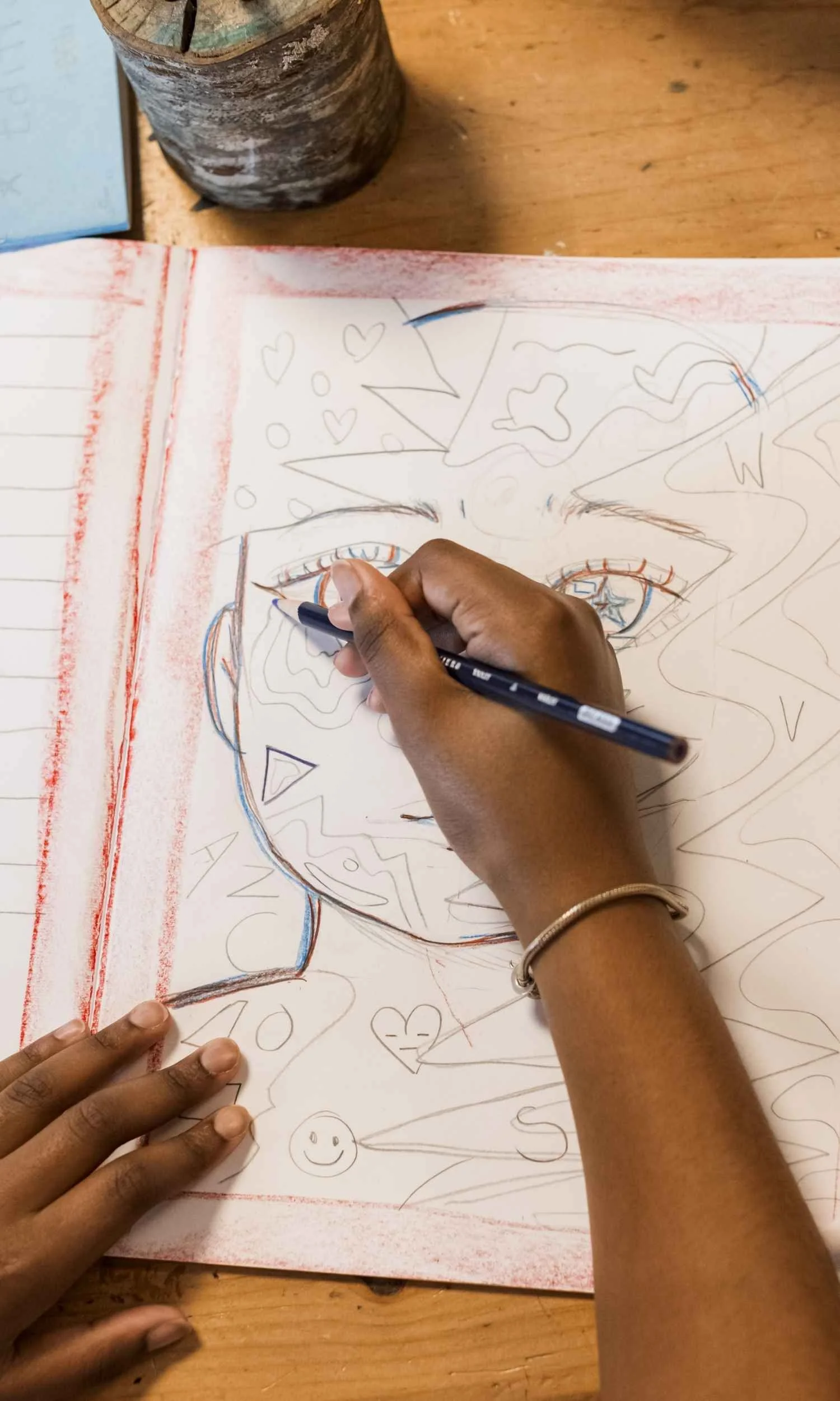 A Nantucket Lighthouse School student draws a colorful abstract portrait in her lesson book, using expressive shapes, patterns, and detailed illustrations as part of a creative classroom learning activity.