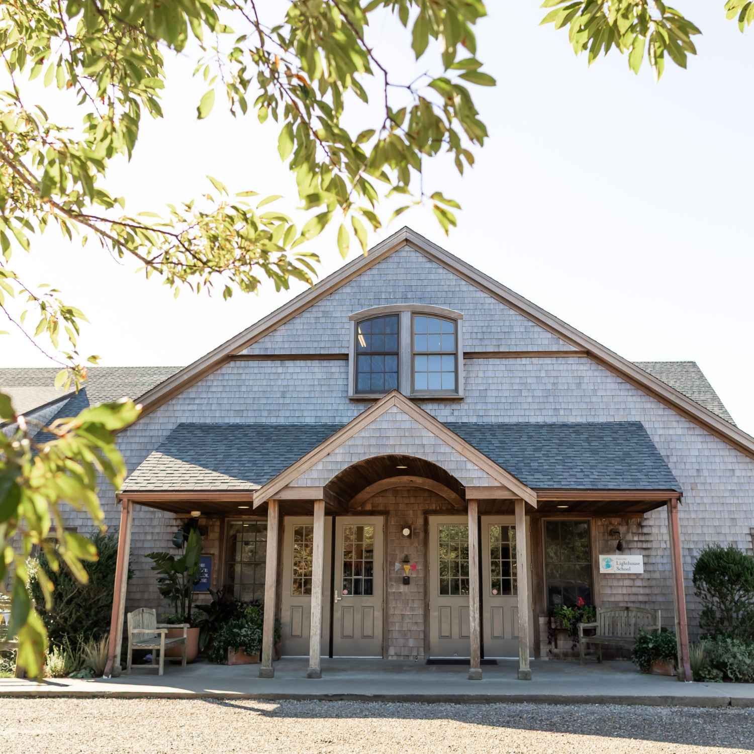 Exterior of the Chace Schoolhouse at Nantucket Lighthouse School, located at 1 Rugged Road on Nantucket, featuring a traditional shingled building with a covered porch, wooden columns, a gabled roof, and a large arched window surrounded by greenery.