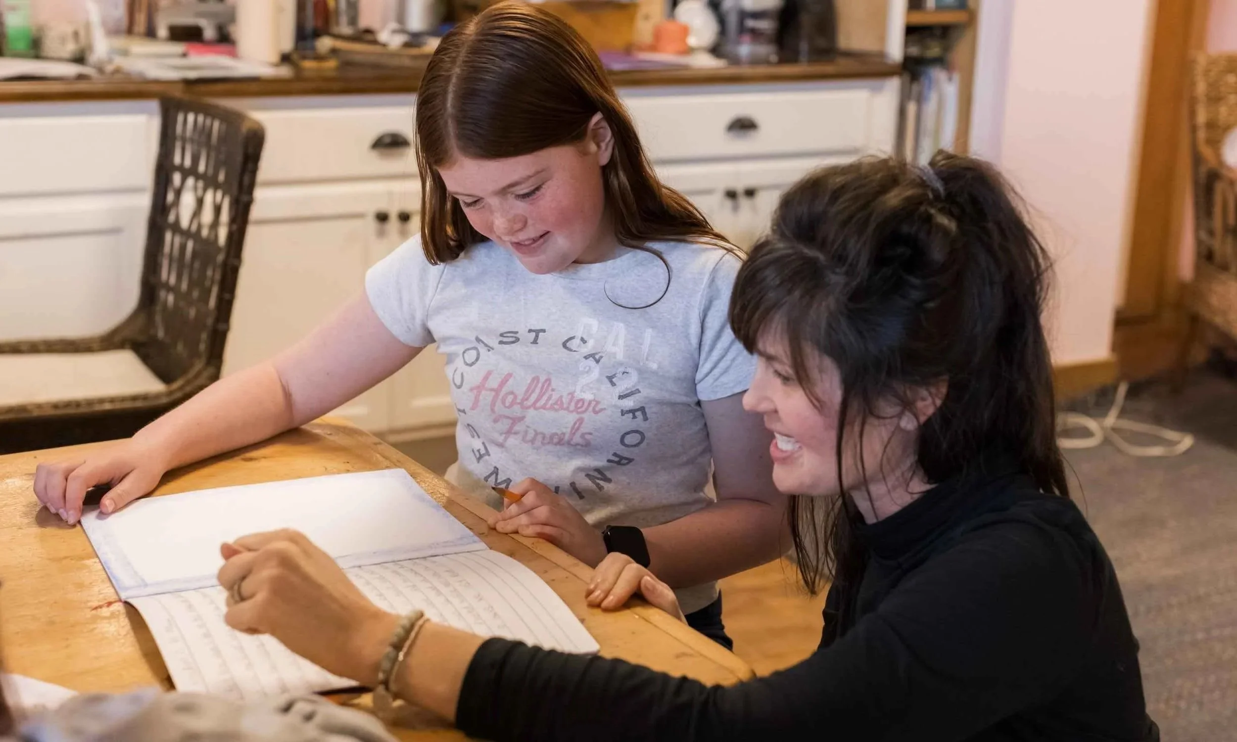 Caitlin Tyler, Nantucket Lighthouse School’s 5th and 6th Grade Teacher and Head of Upper Grades, shares a joyful moment with a Middle School student as they look through a notebook together at a wooden classroom table, with bookshelves in the backgro