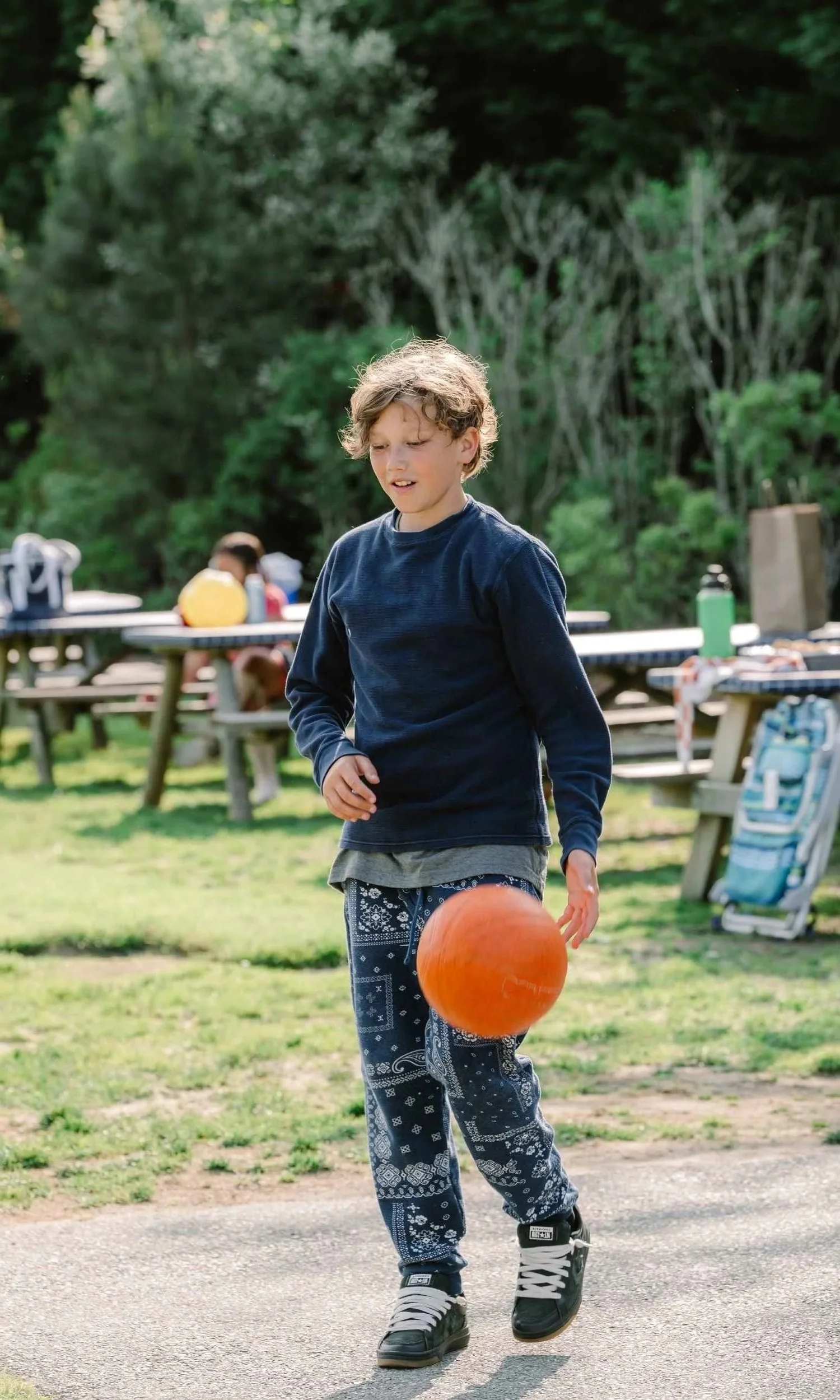 A Nantucket Lighthouse School Middle School student plays basketball outdoors, dribbling an orange basketball near picnic tables and trees while enjoying physical activity and time outside.