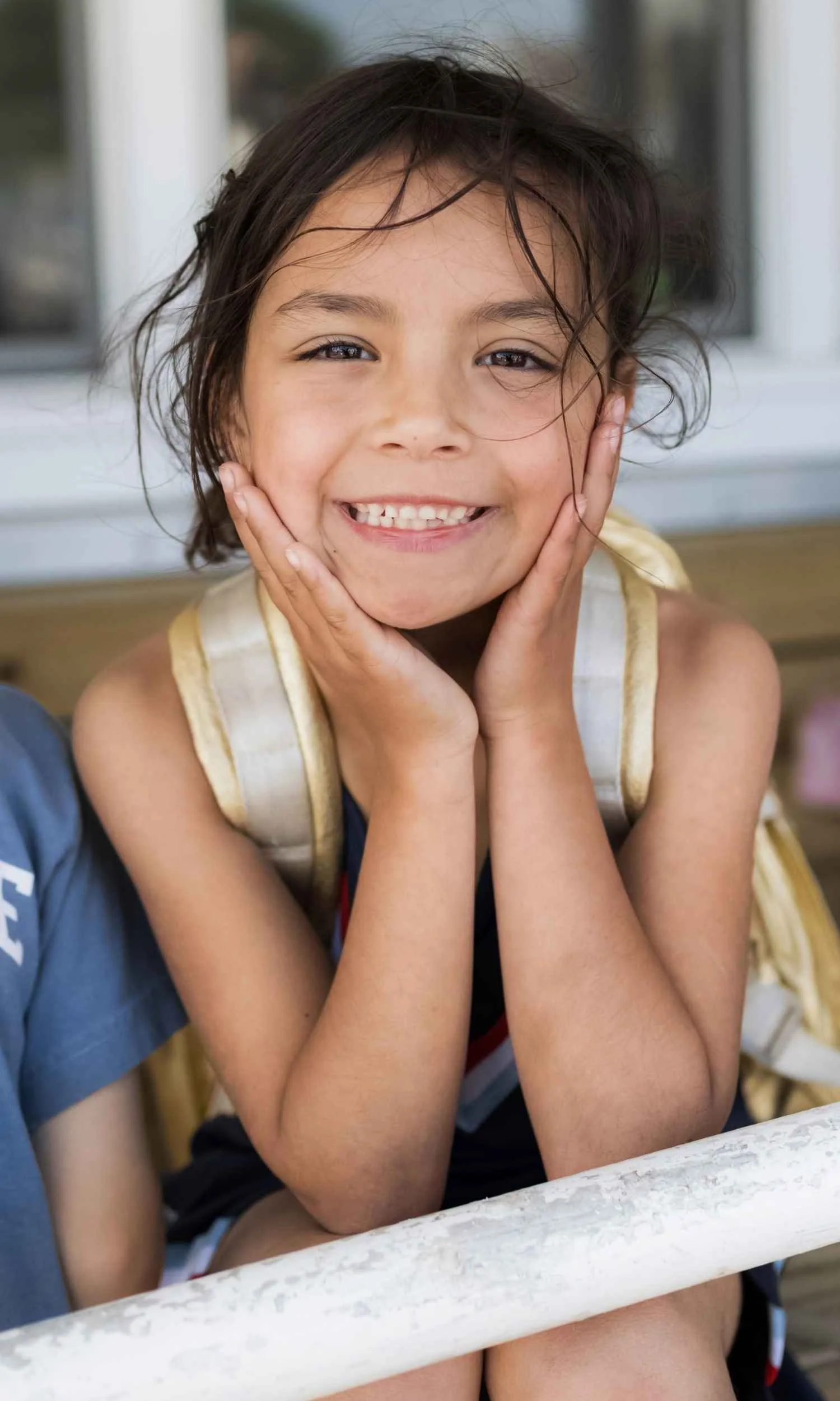 A Nantucket Lighthouse School Student with curly brown hair smiling and holding her face with both hands, sitting outdoors near a white railing.
