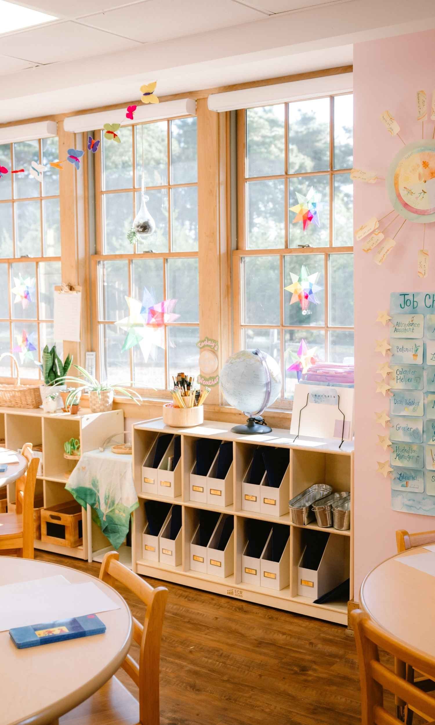 A Nantucket Lighthouse School classroom with large windows decorated with hanging paper butterflies and stars, a globe on a wooden shelf, and a pink wall with educational charts. Wooden furniture and storage bins are visible.