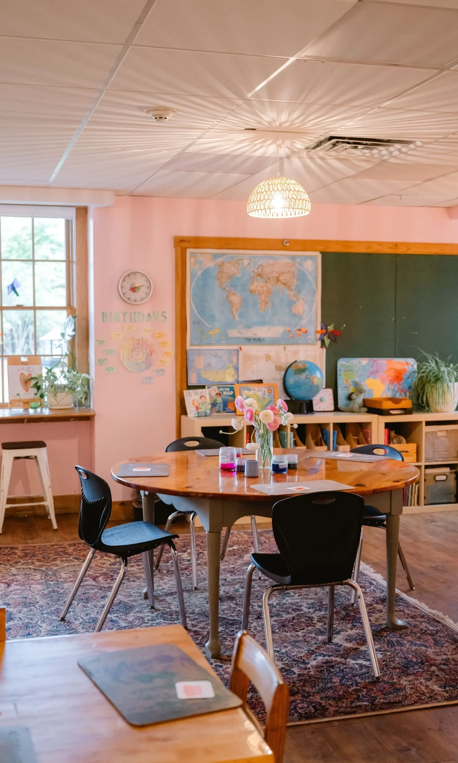 A bright Nantucket Lighthouse School classroom featuring wooden tables and chairs, world maps, books, and plants, with soft pink walls and large windows that fill the welcoming learning space with natural light.