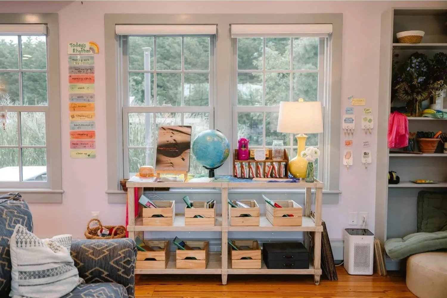 A welcoming learning space at Nantucket Lighthouse School featuring wooden shelves filled with books, storage bins, a globe, and a table lamp, with large windows overlooking the surrounding trees.