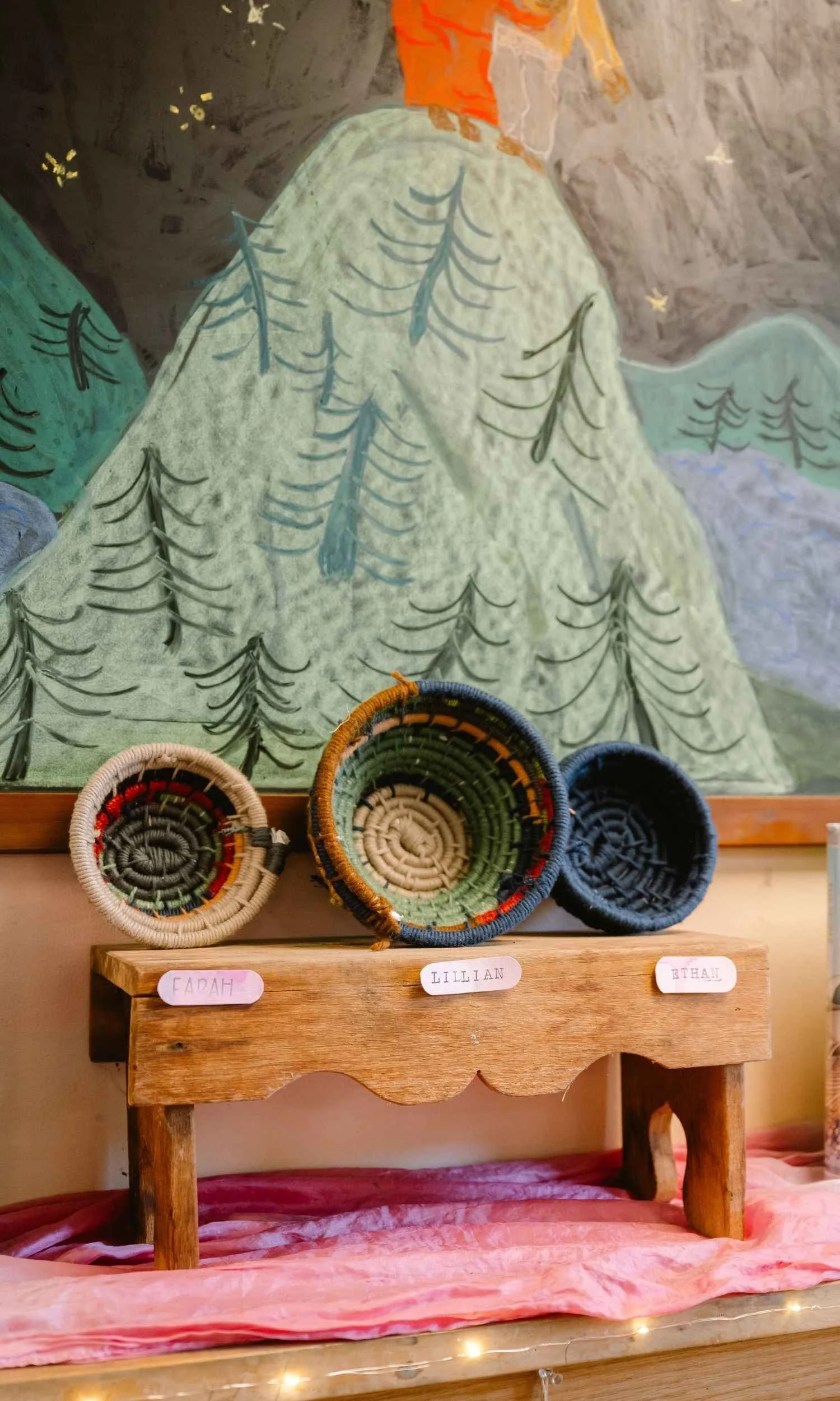 Colorful woven baskets displayed on a small wooden table, with labels reading 'Fadah,' 'Lillian,' and 'Ethan.' In the background, a mural depicts a mountain landscape with trees, stars, and an orange sun or sunset.