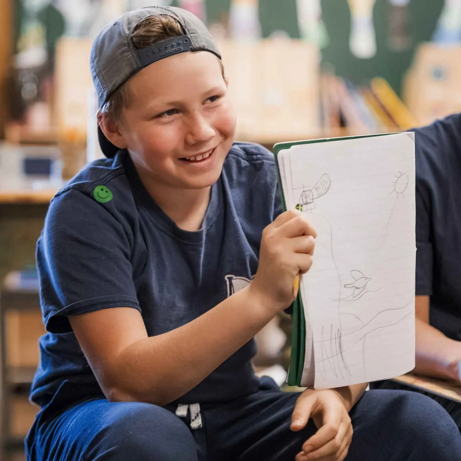 A smiling Nantucket Lighthouse School student wearing a backwards cap, holding a notebook with drawings of a helicopter, a fish, and a sun, in a classroom setting.