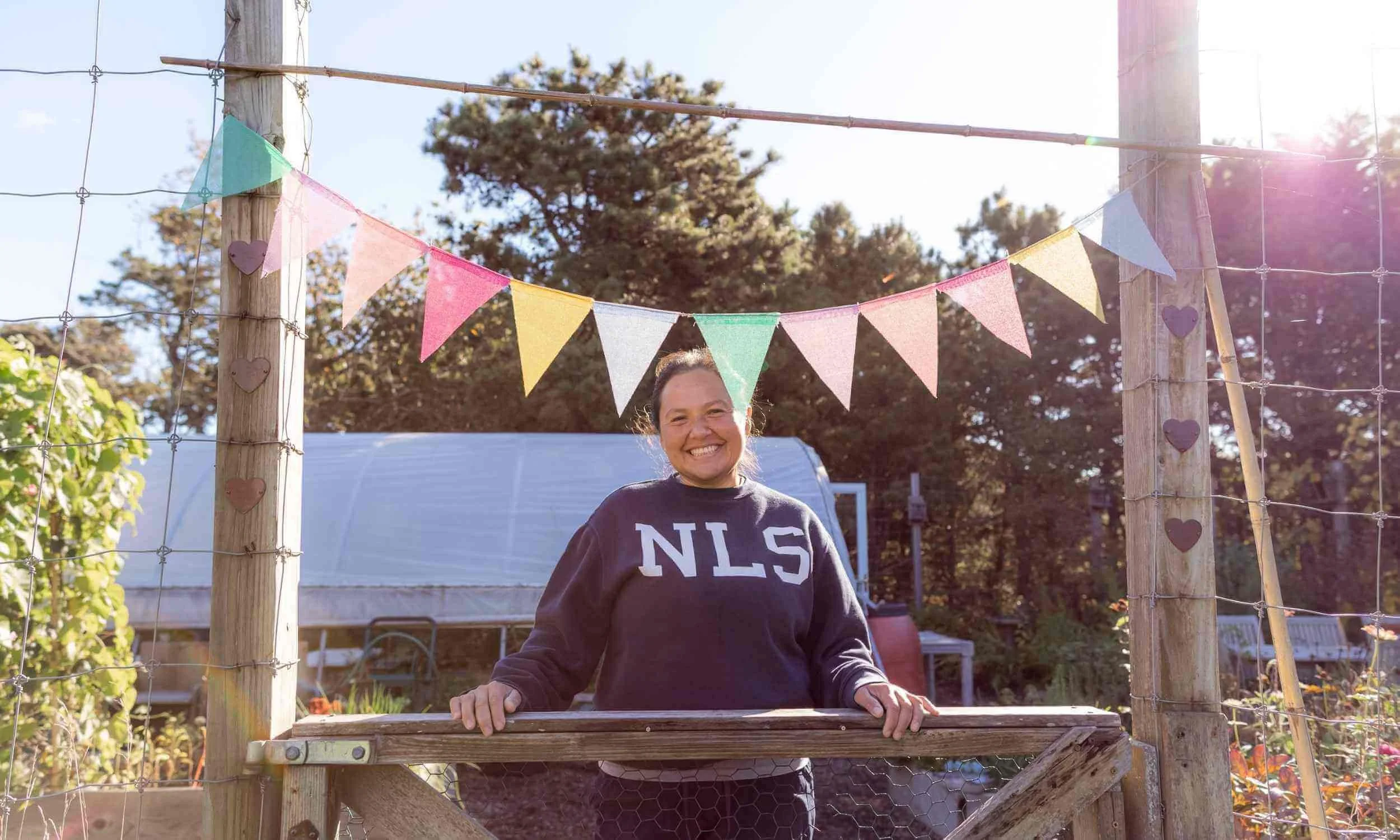 Nantucket Lighthouse School’s horticulture teacher smiles while standing at the entrance to the school garden, surrounded by trees and sunlight, with a colorful pennant flag banner hanging above the wooden gate.