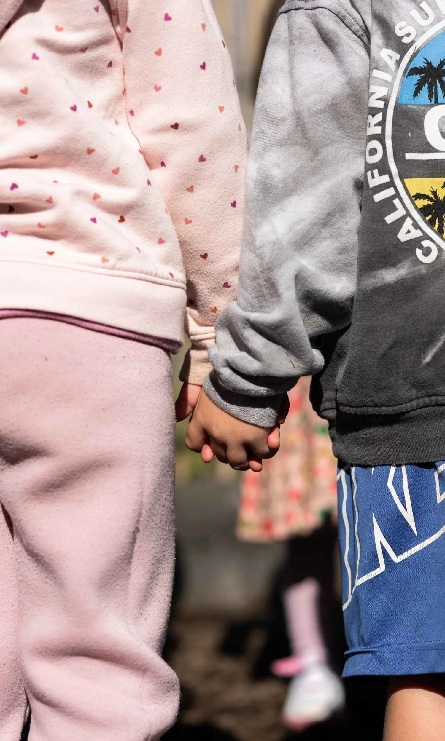 Two Nantucket Lighthouse School Small School students holding hands, one wearing a pink sweatshirt with small heart patterns, the other in a gray sweatshirt and blue shorts.