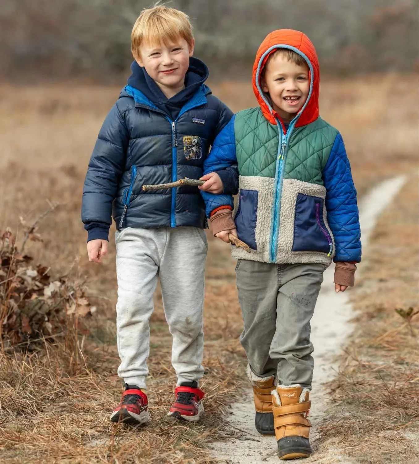 Two Nantucket Lighthouse School Kinderclass students explore the outdoors together, walking along a wooded path while carrying sticks and dressed in warm jackets and boots.