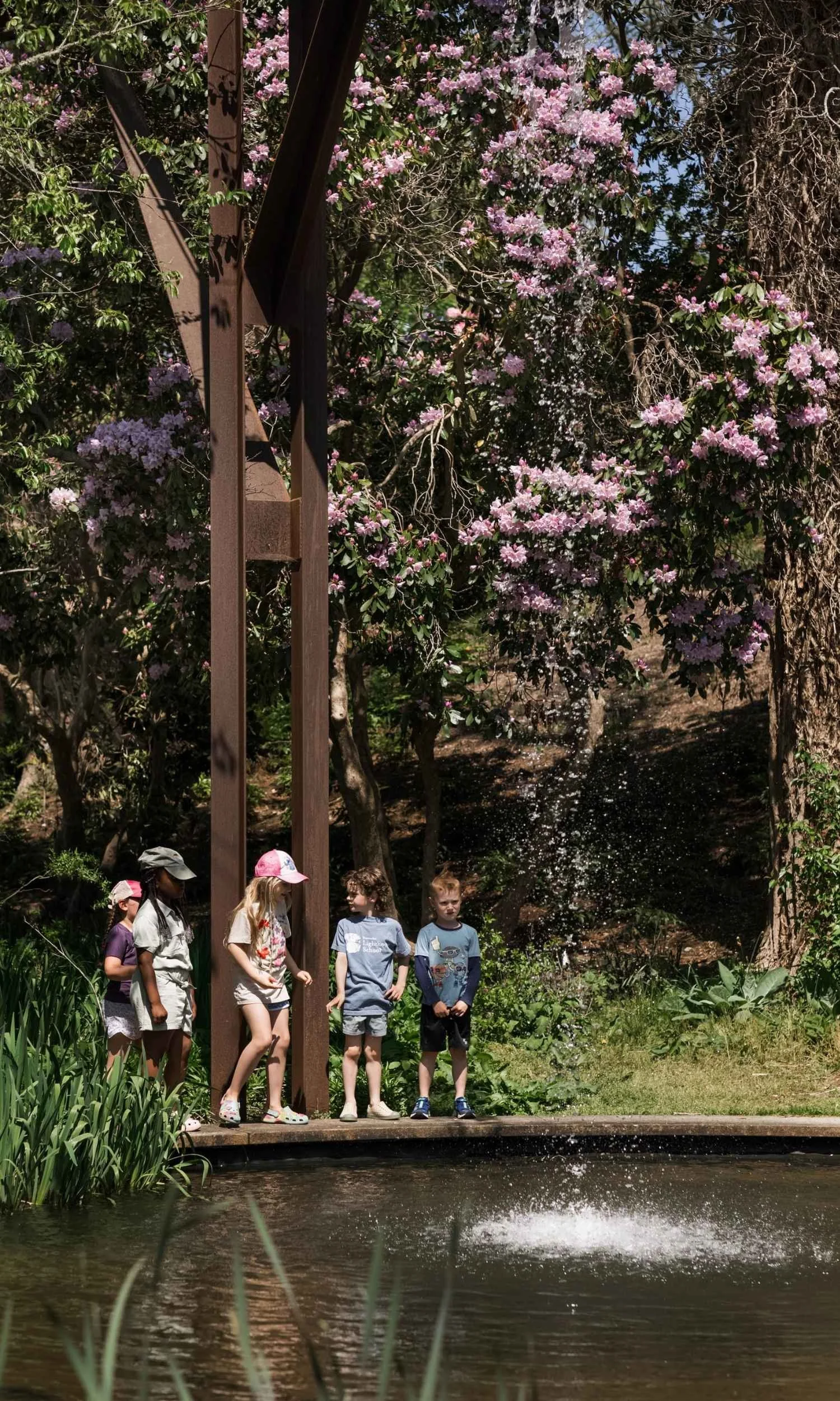 A group of Nantucket Lighthouse School students standing on a wooden deck near a pond with water splashing, surrounded by greenery and pink flowering bushes, with a large brown metal structure and some rocks in the background.