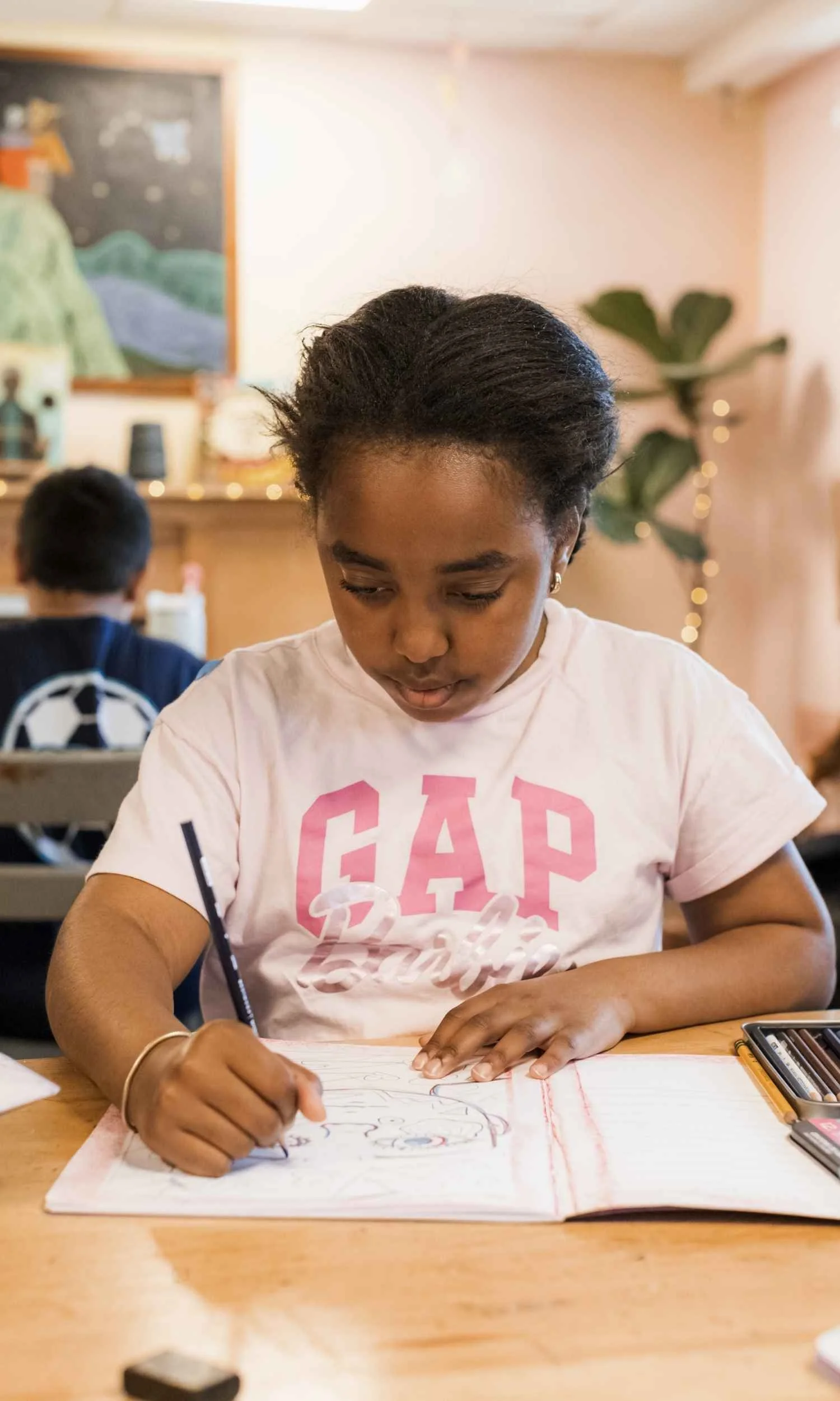 A Nantucket Lighthouse School student sits at a wooden classroom table, focused on drawing, while another student works in the background of the warm, inviting learning space.