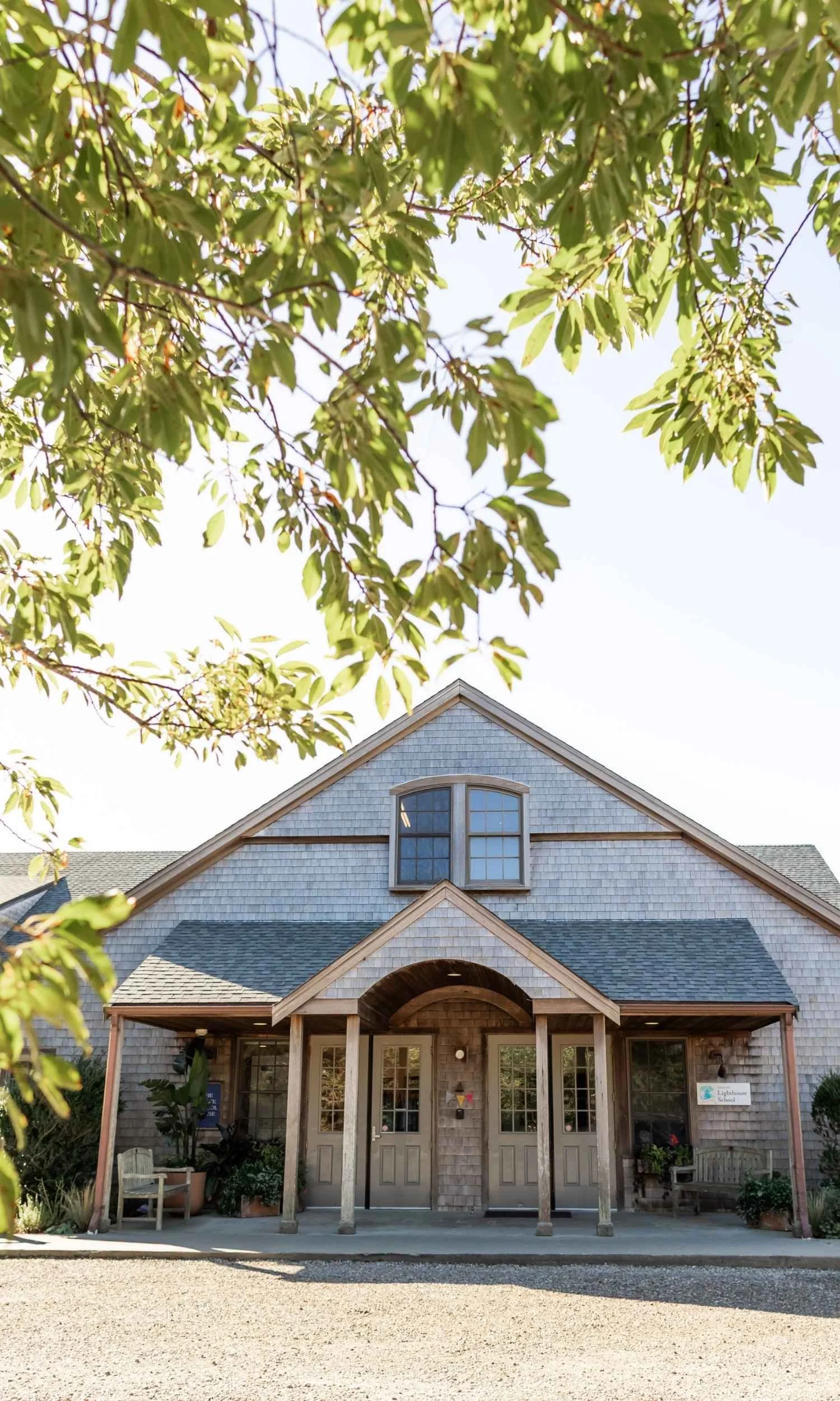 Front view of Nantucket Lighthouse School's Chace School House at 1 Rugged Road with a shingle exterior, timber porch, and a gabled roof, partly obscured by tree branches in the foreground, under a clear sky.