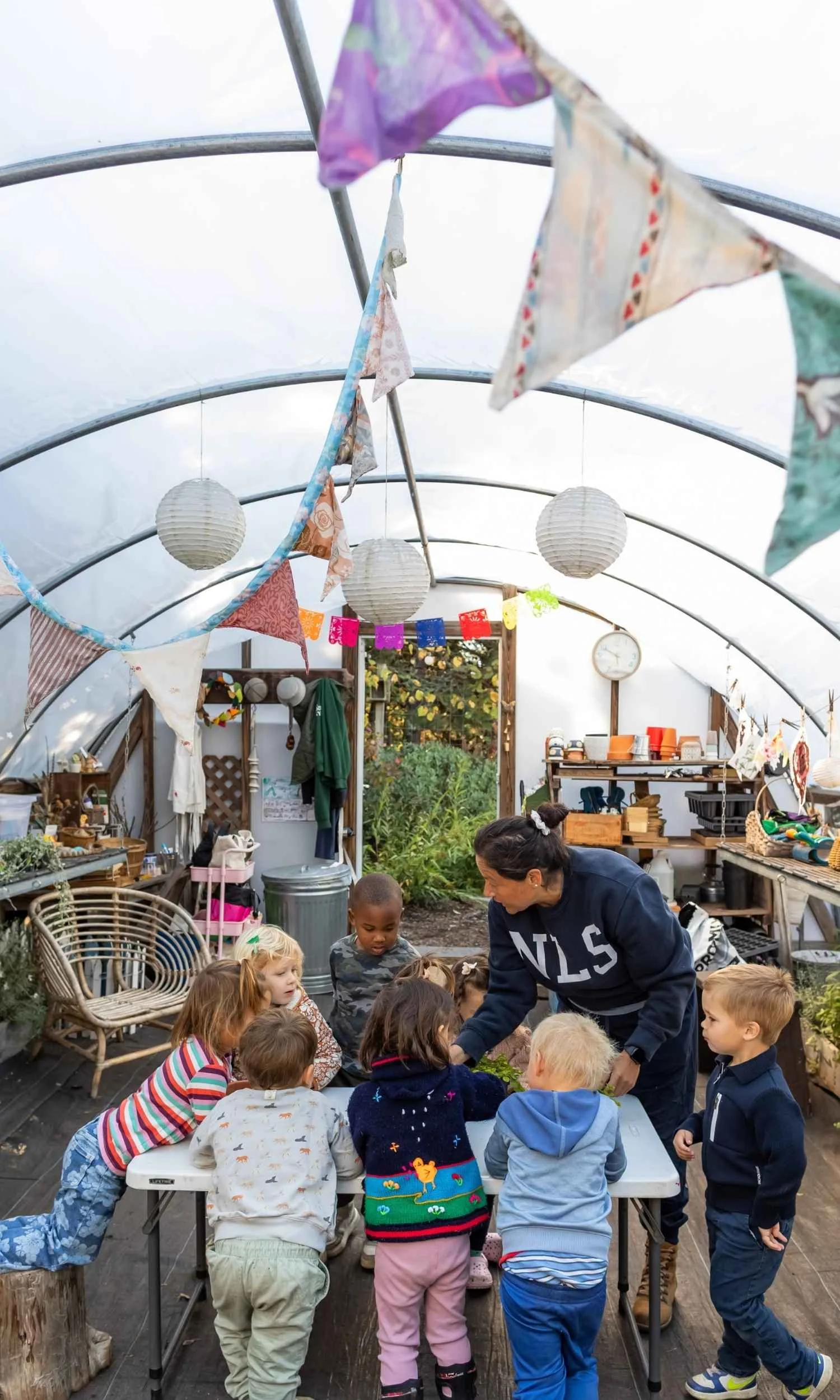 Nantucket Lighthouse School Small School students participate in a hands-on horticulture class with their teacher inside a greenhouse, surrounded by plants, gardening supplies, and colorful decorations.