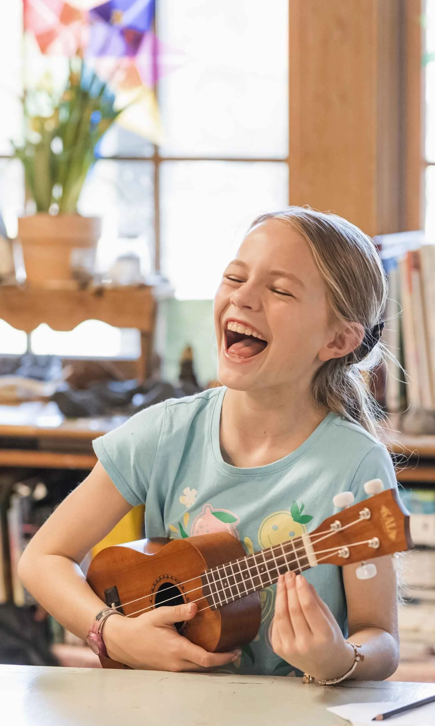 A Nantucket Lighthouse School student with blonde hair tied back, smiling and singing while playing a small wooden ukulele, sitting at a table in a cozy classroom with books, plants, and colorful decorations.