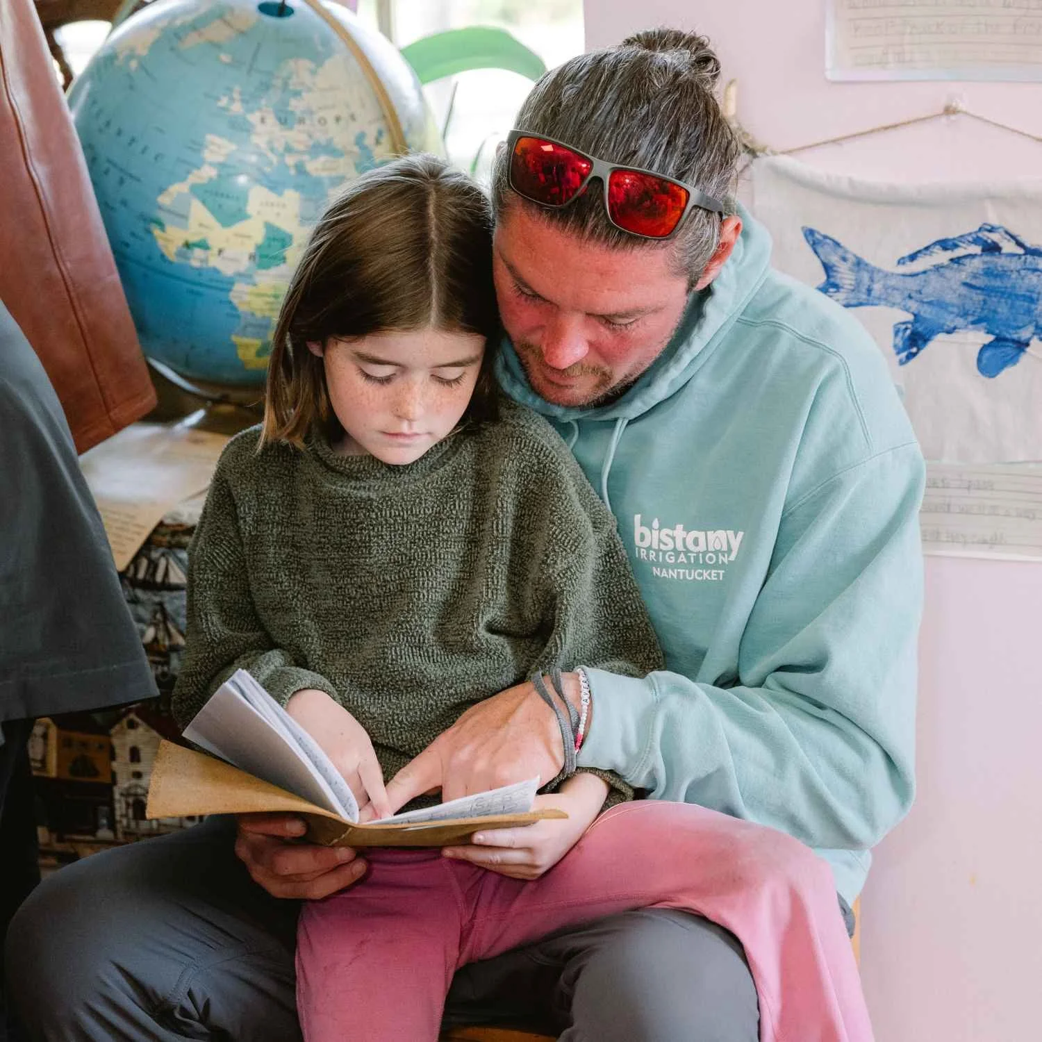 A Nantucket Lighthouse School student and her father share a moment reading together during Project Night, surrounded by student artwork and classroom displays celebrating learning and creativity.