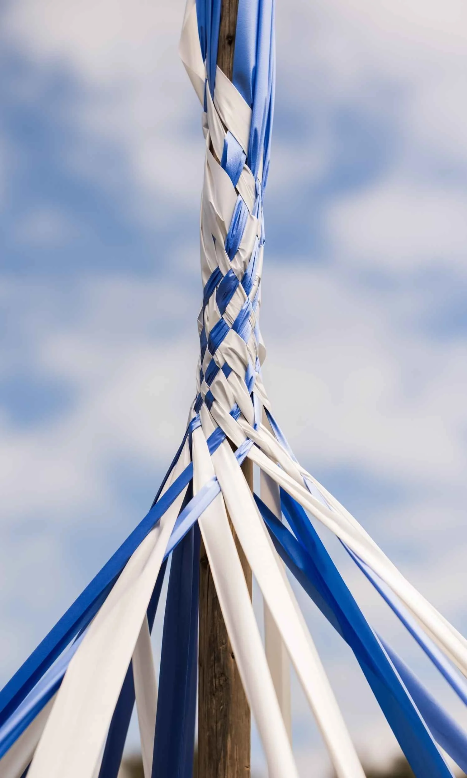 A traditional Maypole at Nantucket Lighthouse School, decorated with intricately woven blue, white, and light blue ribbons extending from the wooden pole, celebrating the school's annual spring Maypole tradition.