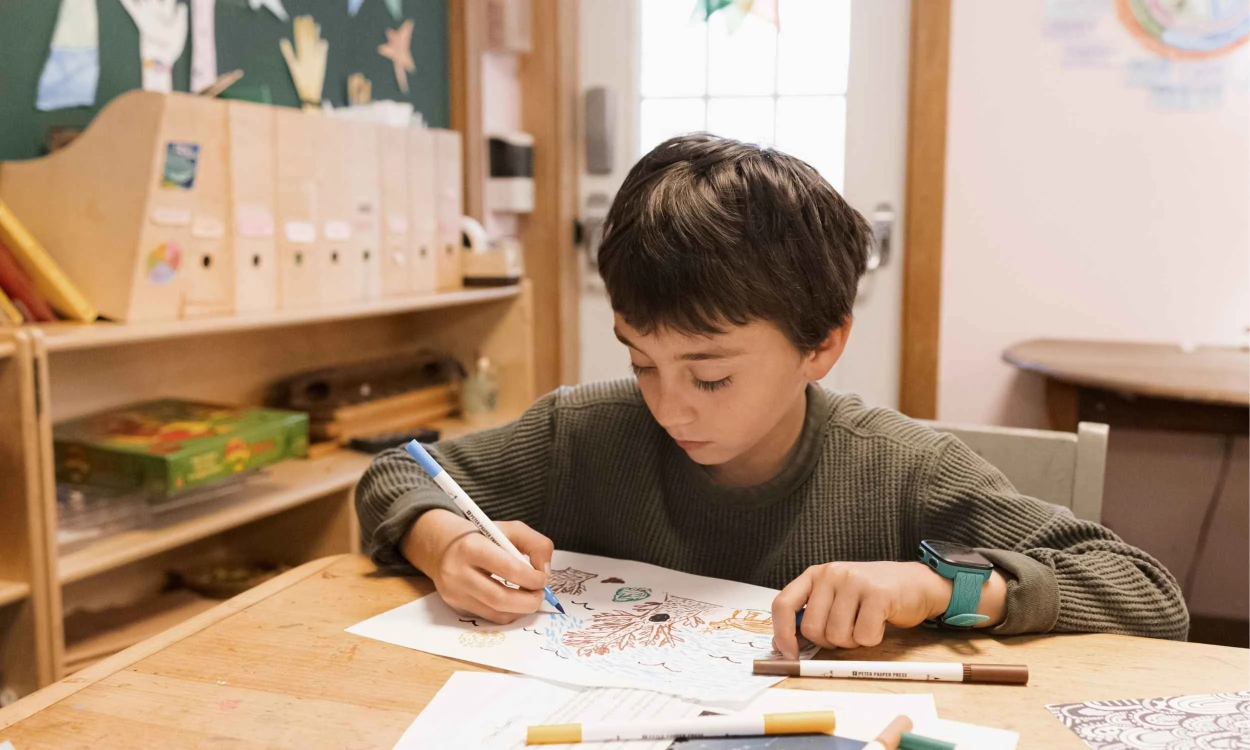 A Nantucket Lighthouse School student with brown hair sitting at a wooden table, drawing or coloring on a sheet of paper with markers. The background features a classroom with educational posters and storage shelves.