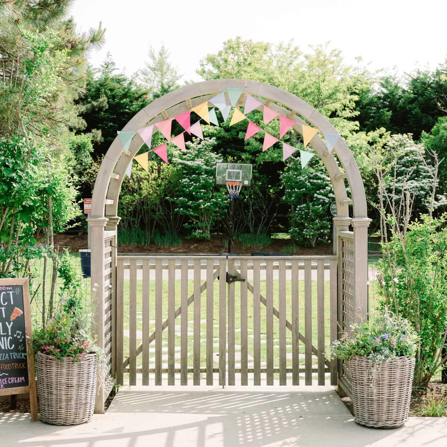 A garden gate at Nantucket Lighthouse School, decorated with a colorful pastel pennant flag banner and surrounded by potted plants, flowers, and lush greenery, with a basketball hoop visible in the background.