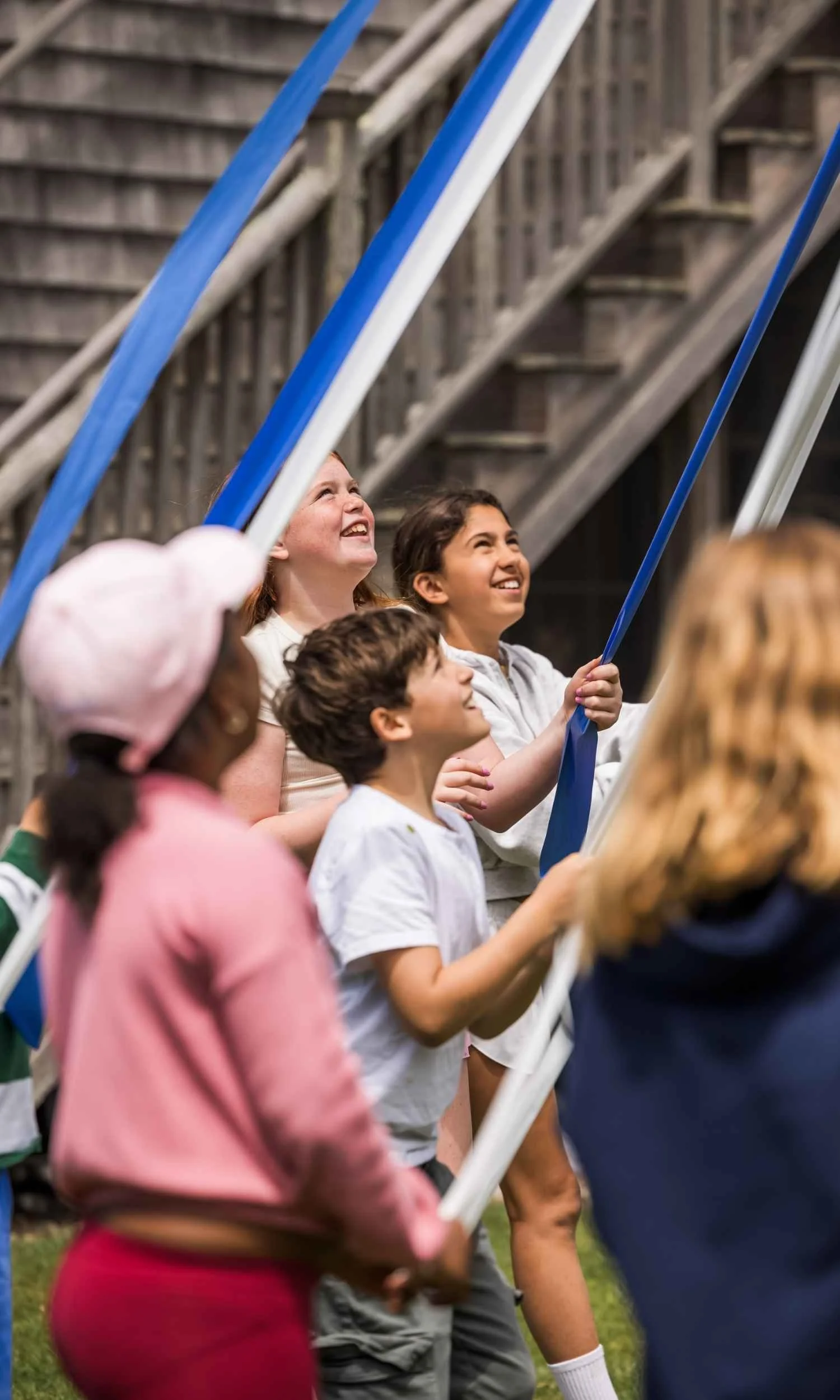 Nantucket Lighthouse School students participate in the traditional Maypole celebration, joyfully holding blue and white ribbons during an outdoor spring gathering surrounded by trees and nature.