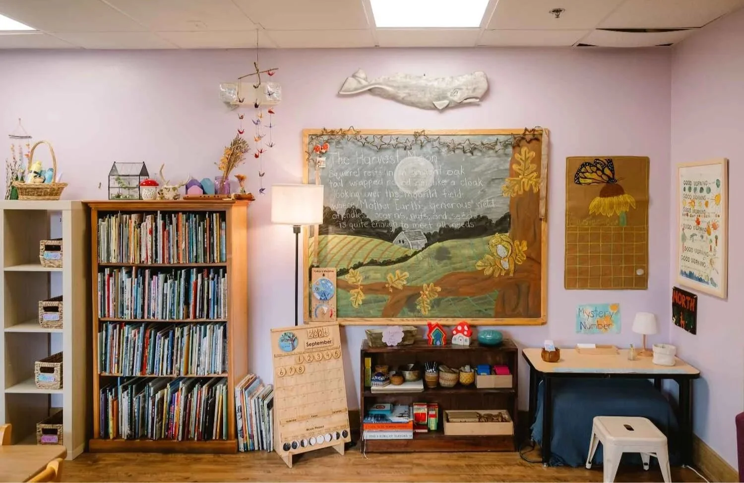 A Nantucket Lighthouse School classroom corner featuring a bookshelf filled with children's books, art and craft supplies, educational posters, and a chalkboard with handwritten lessons and colorful illustrations.