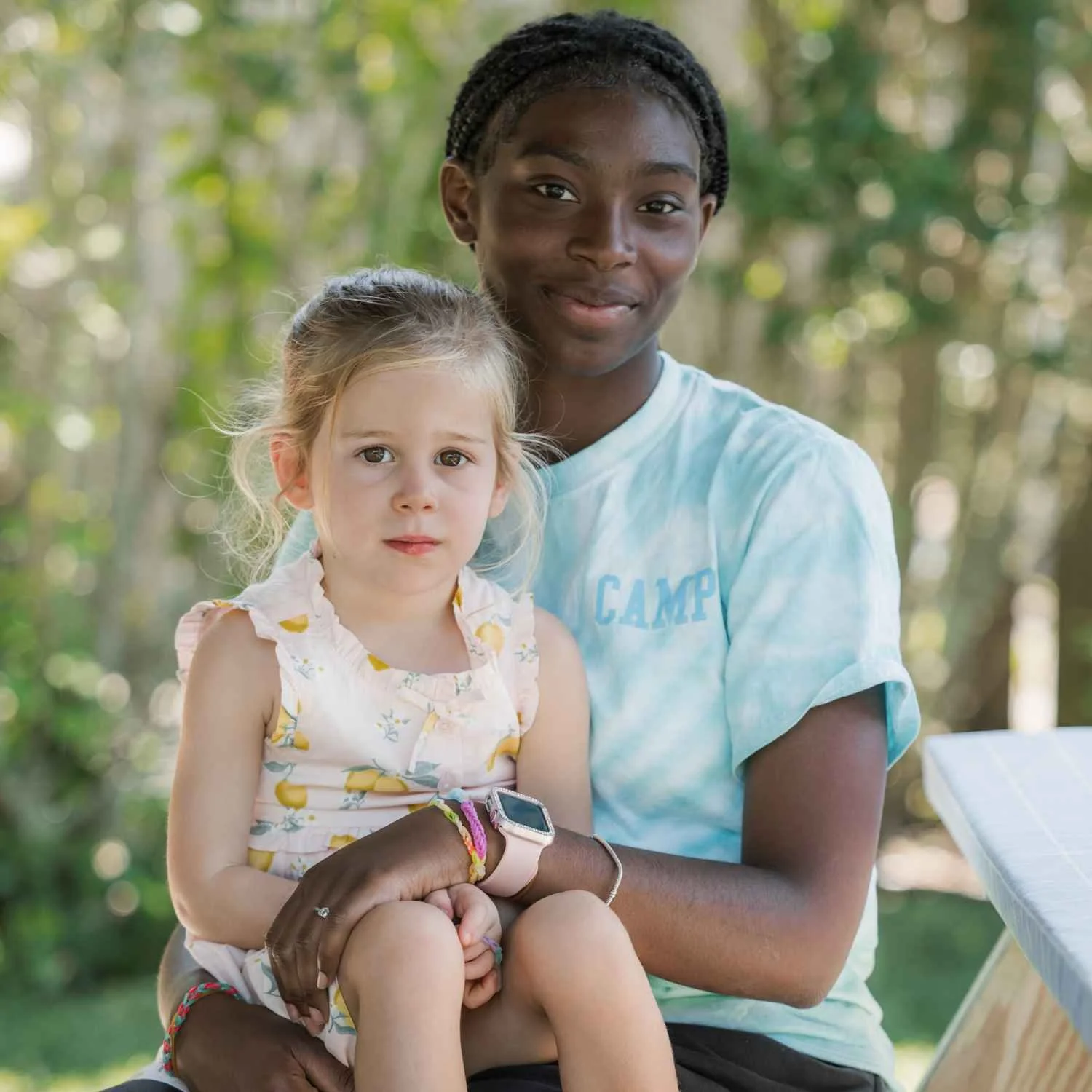 A Nantucket Lighthouse School Camp Lighthouse counselor holds a young camper outdoors in a wooded setting, surrounded by trees and nature, as they pose together for the camera.