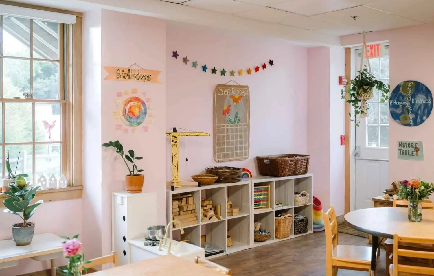 A bright Nantucket Lighthouse School classroom featuring wooden furniture, colorful decorations, a nature table, indoor plants, and organized learning materials, with sunlight streaming through large windows and fresh flowers on a round table.
